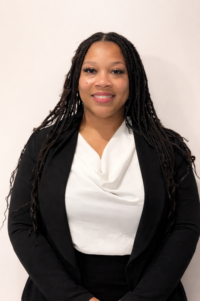 A smiling woman with long braided hair, wearing a black blazer and a white blouse, standing against a plain white background.