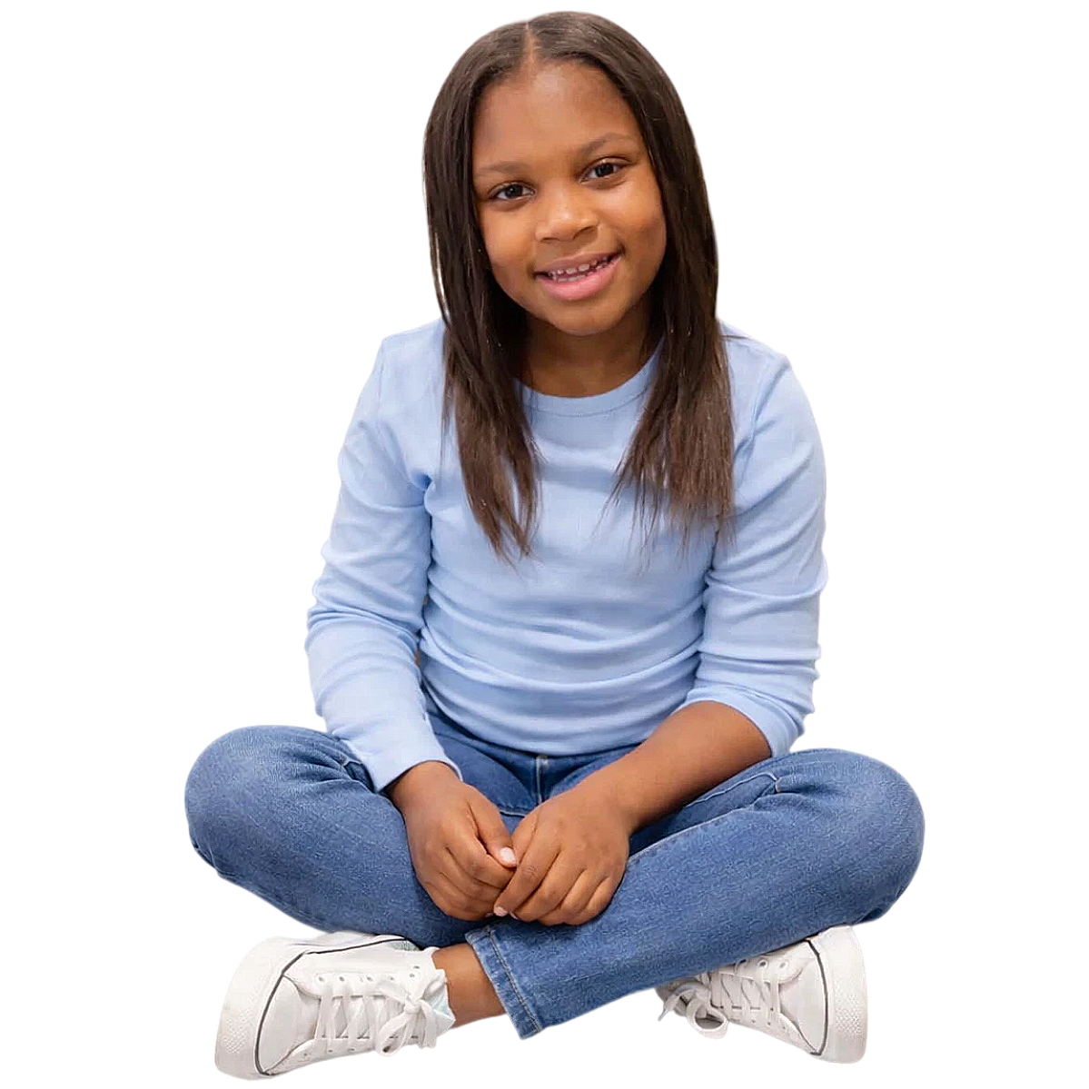 A girl with long dark hair sitting cross-legged on the floor, wearing a light blue long-sleeve shirt, blue jeans, and white sneakers, smiling at the camera.