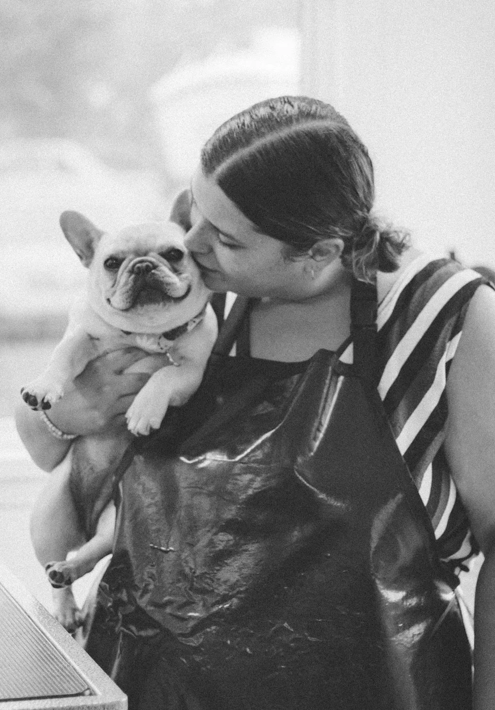 A woman with her hair in a braid holds a French Bulldog close and kisses it.