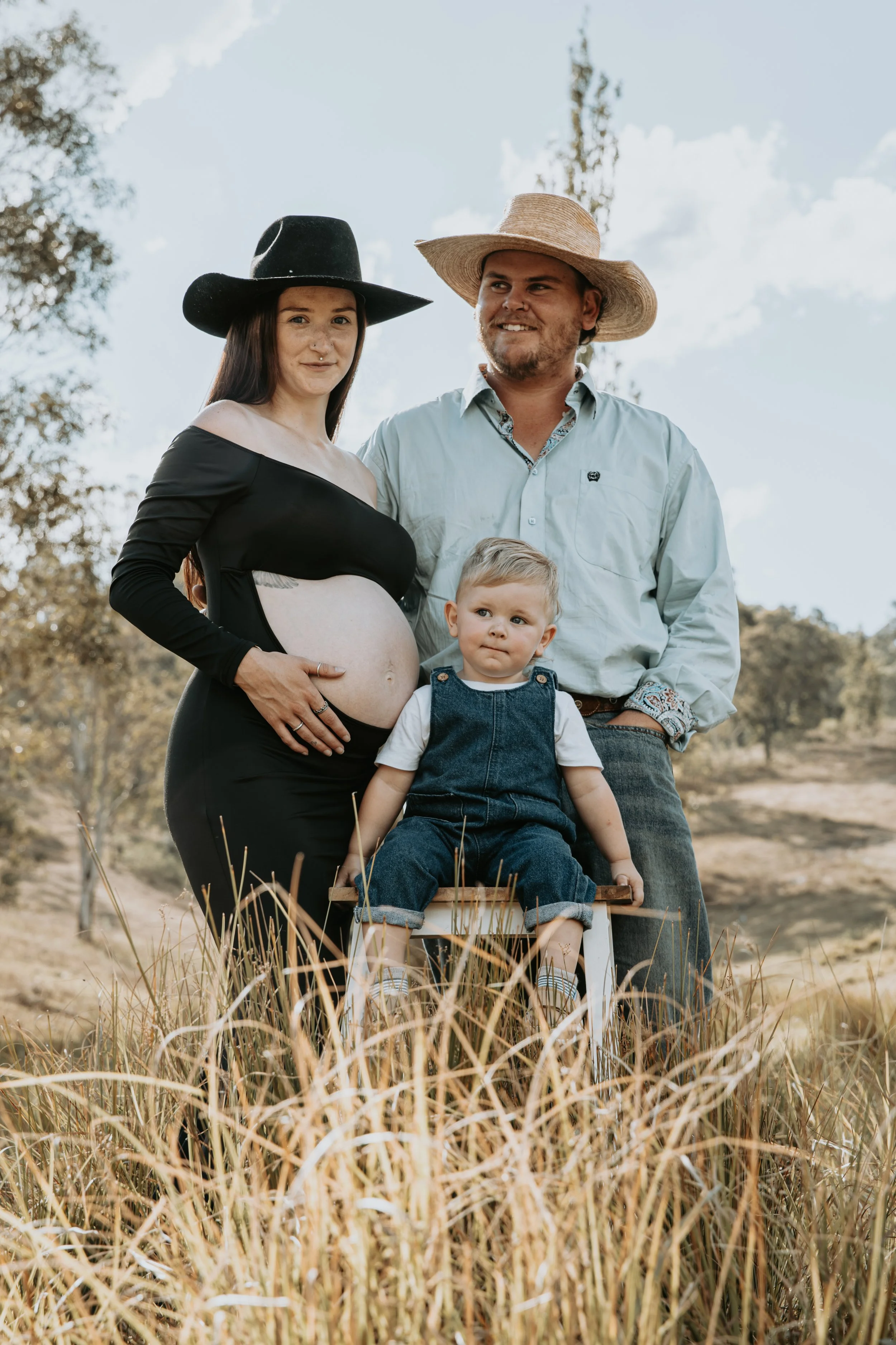 A pregnant woman, a man, and a child are outdoors in a grassy field with trees in the background. The woman is wearing a black wide-brimmed hat and a black dress, and the man is wearing a straw hat and a light blue shirt. The child, sitting on a smal