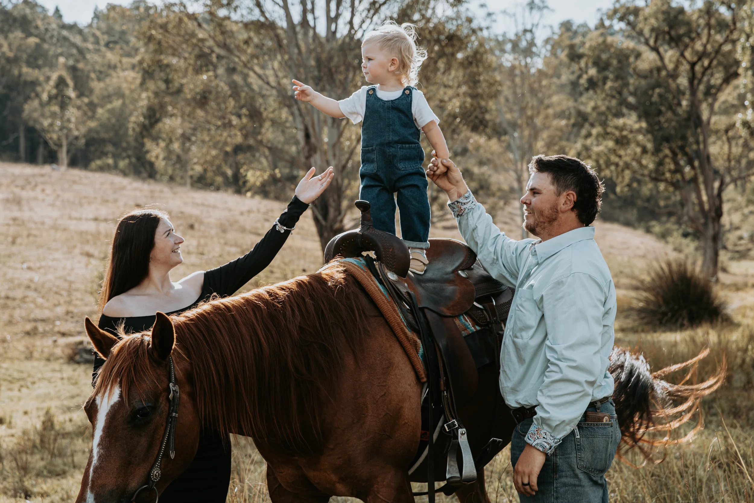 A family outdoors with a young girl standing on a saddle on a horse, supported by a man, while a woman reaches out to her.