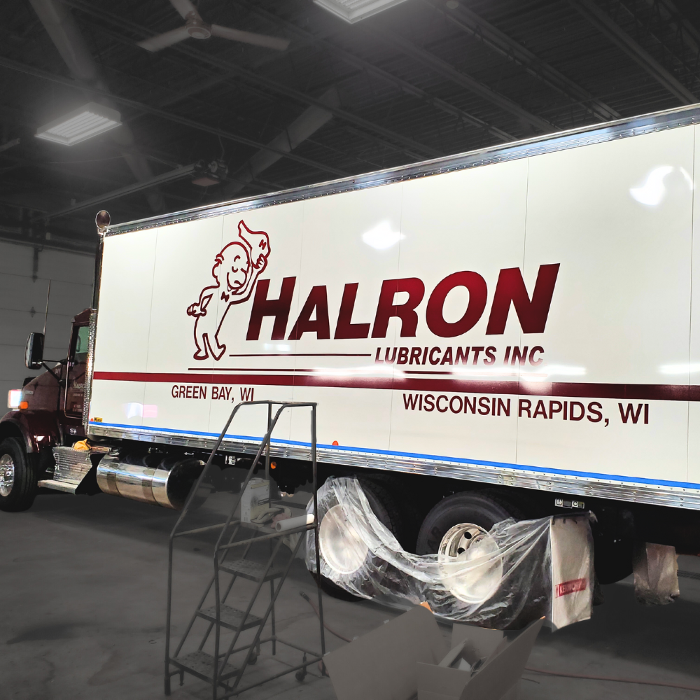 A large truck inside a workshop with scaffolding and painter's plastic covering the wheels, displaying the logo of Halron Lubricants Inc. with a smiling cartoon character and text indicating locations in Wisconsin.