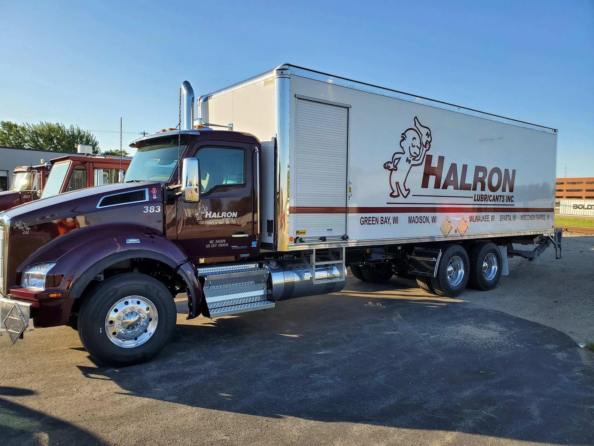 A large semi-truck with a dark purple cab and tan trailer featuring a lion mascot and the words HALRON LUBRICANTS INC. with locations listed below, parked on a gravel lot in the sunlight.