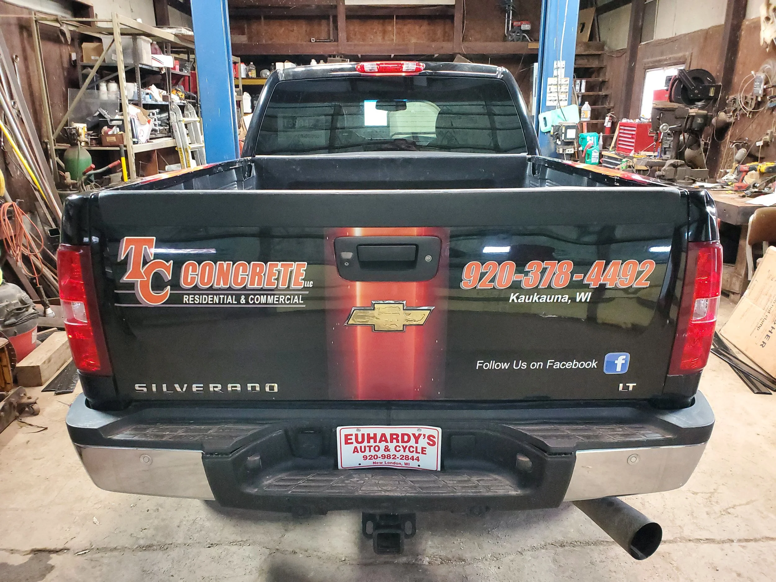Black Chevrolet Silverado pickup truck with advertising decals for TC Concrete and Euhardy's Auto & Cycle on the tailgate, parked inside a workshop or garage.