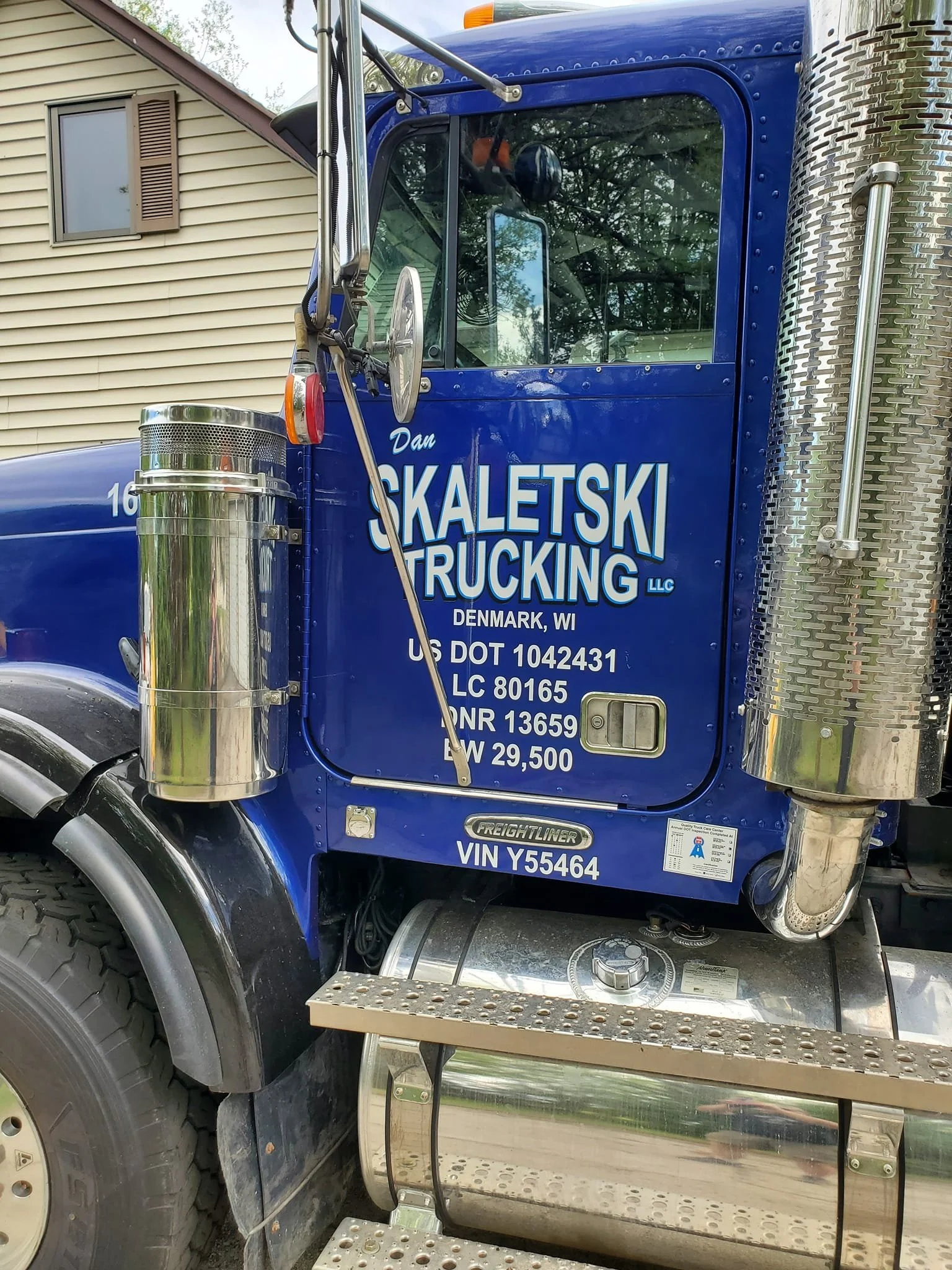 Close-up of a blue semi-truck cab with white lettering that reads 'Dan Skaltski Trucking LLC Denmark, WI' and various licensing numbers. The truck has chrome exhaust pipes, a fuel tank, and metal steps beneath the door. The background shows part of a