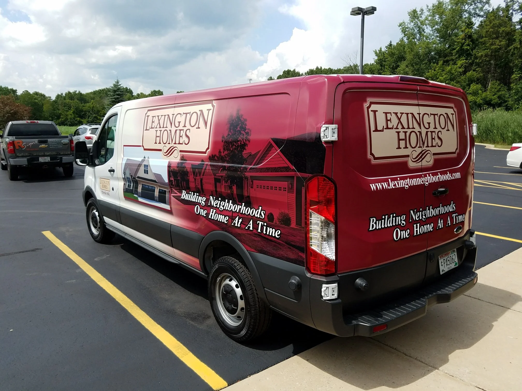 A Lexington Homes branded delivery or service van parked in a lot, with a red and white design showcasing houses and the company logo, and the slogan "Building Neighborhoods One Home At A Time."