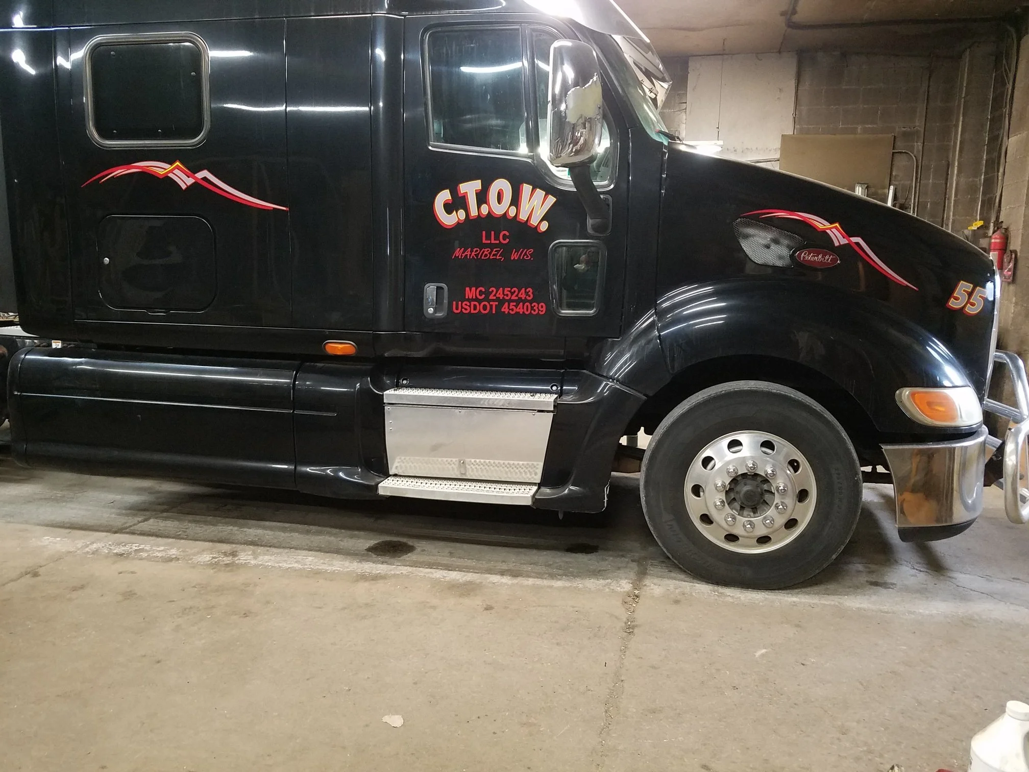 Black semi-truck with red and white decals and lettering on the side parked indoors, with the front of the truck facing right.