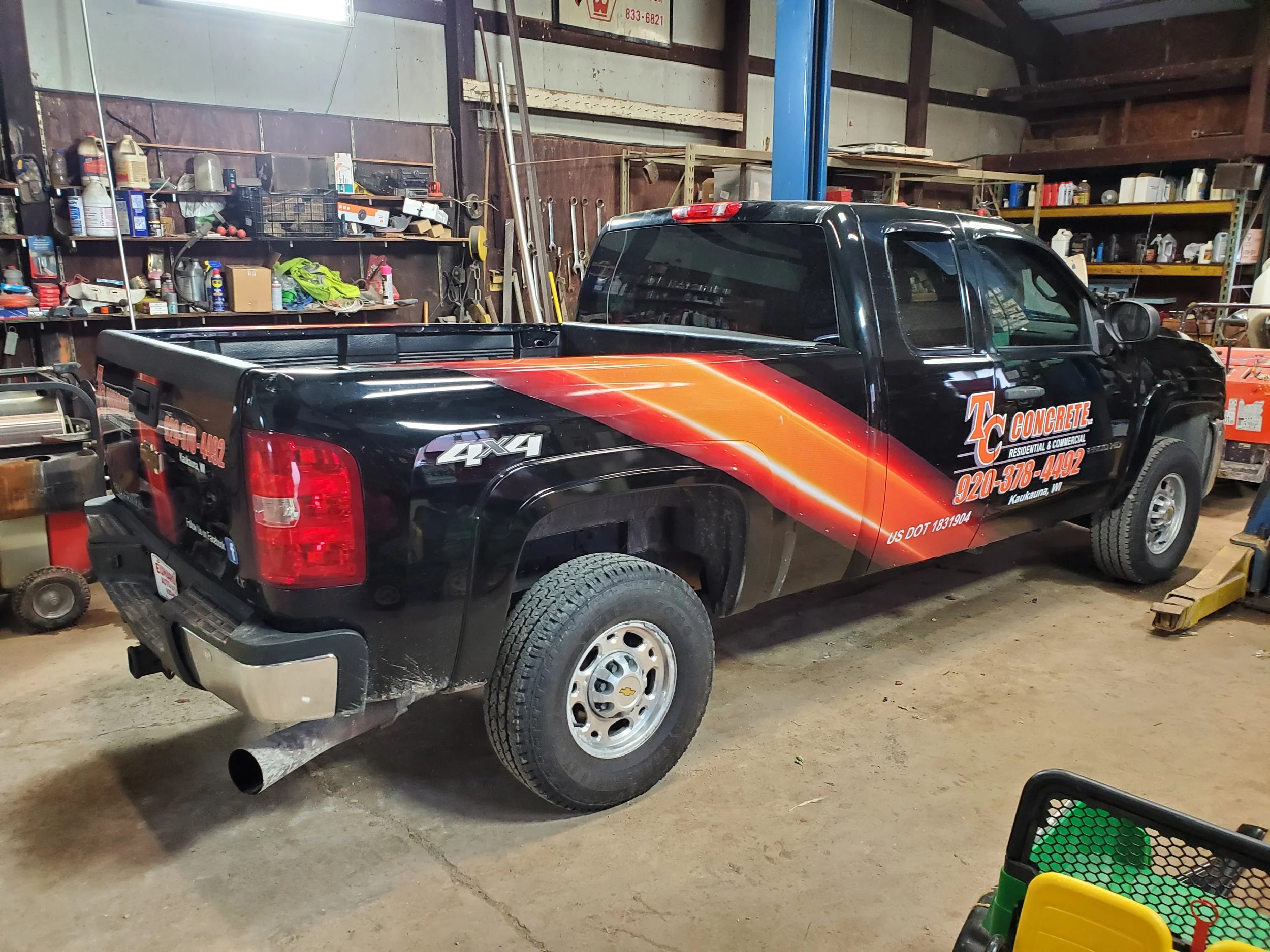 Black pickup truck with orange and red graphics for TC Concrete, parked inside a garage or workshop with tools and equipment on shelves and hanging on the walls.