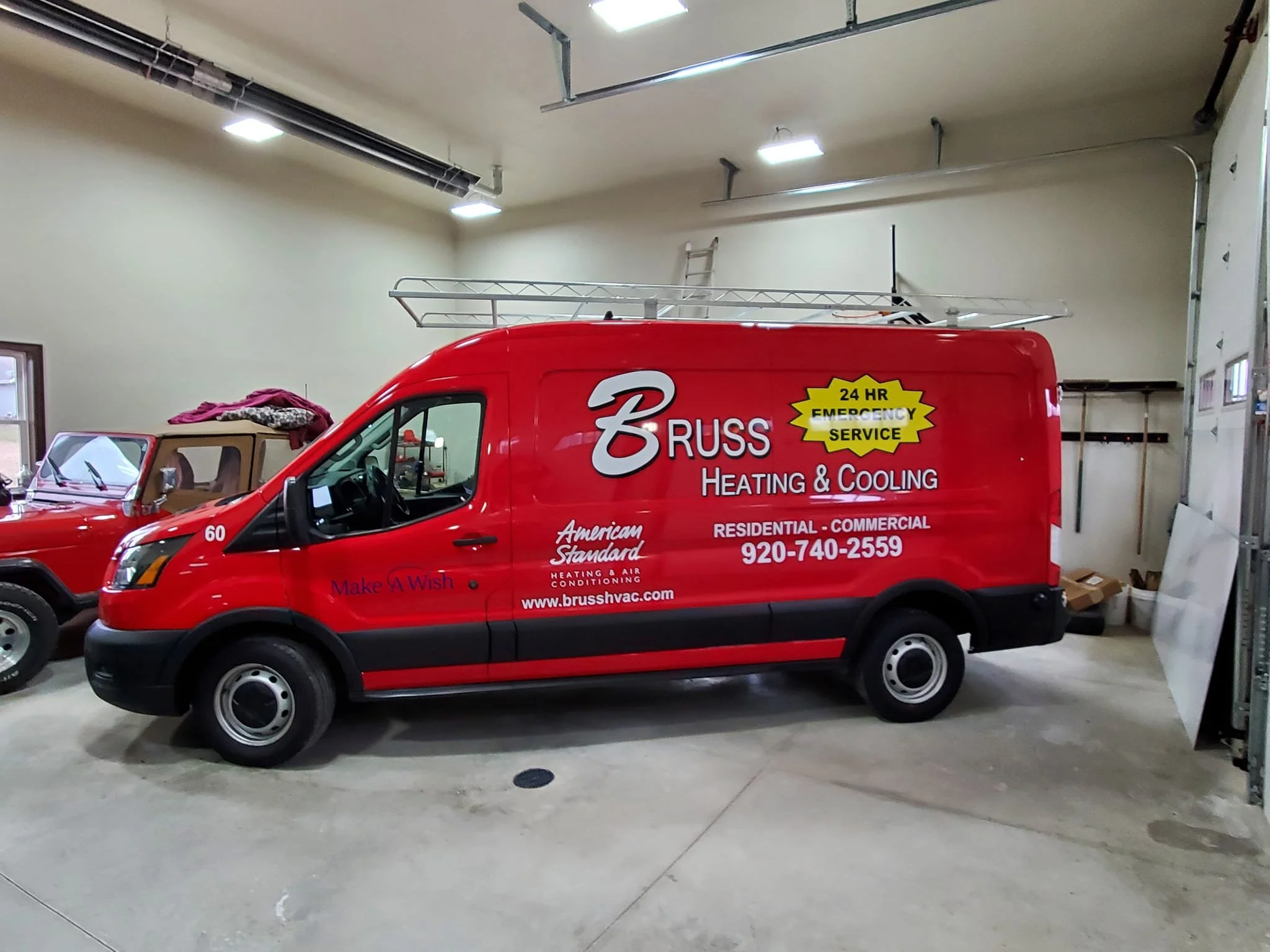 Red service van with company logo and phone number inside a garage, alongside another vintage vehicle.