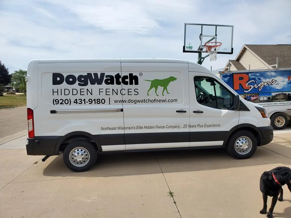 A white DogWatch Hidden Fences service van parked on a driveway with a basketball hoop behind it. A black dog is near the bottom right corner of the image.
