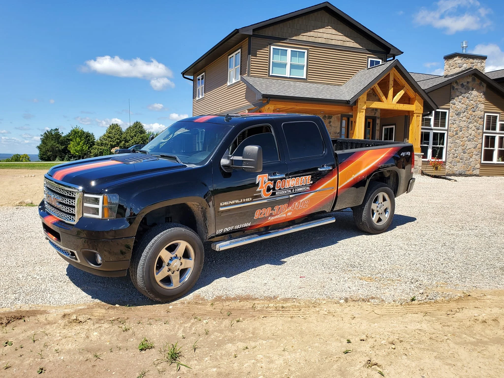 Black GMC pickup truck with orange and red company logo and contact information parked in front of a large house with stone and wood siding, on a gravel driveway under a blue sky.