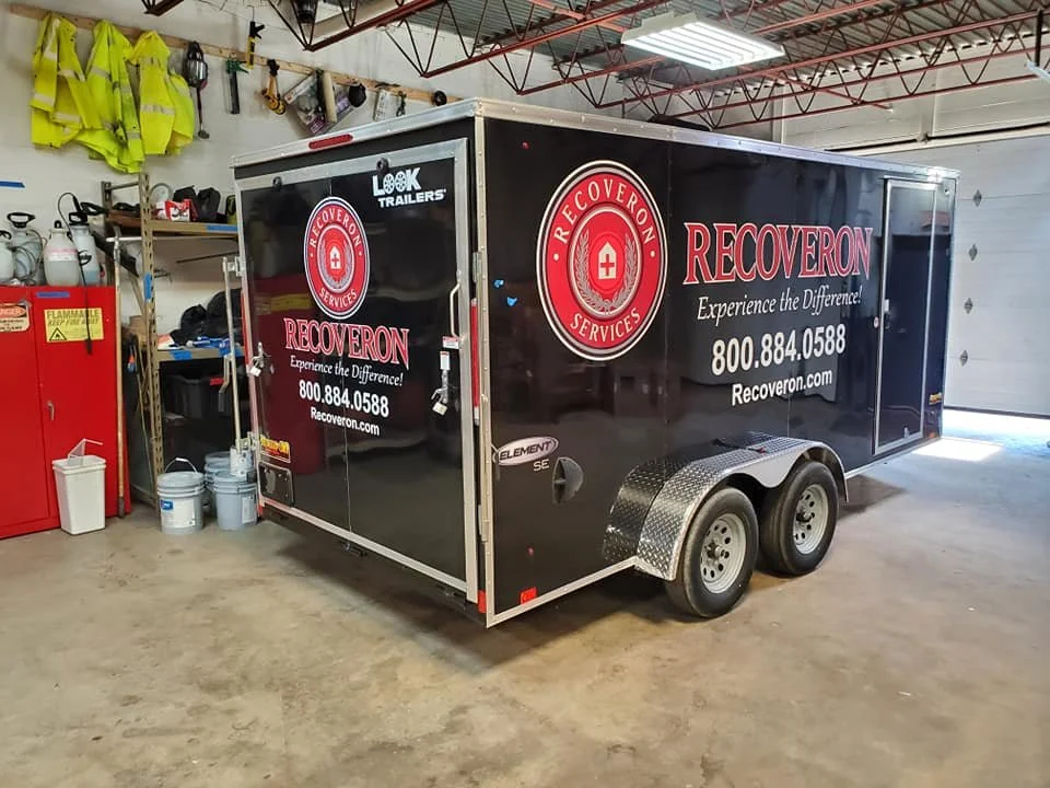 A black enclosed trailer with red and white branding for Recoveron Services, parked inside a garage or workshop. The trailer has dual wheels and safety reflective markings, with equipment and safety gear hanging on the wall in the background.