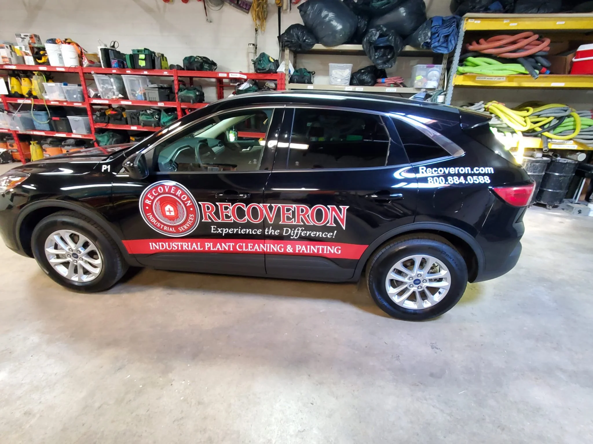 Black company vehicle inside a warehouse with red shelving, containing tools and equipment, advertising 'Recoveron' industrial plant cleaning and painting services, with the website recoveron.com and phone number 800.884.0588 on the rear side.