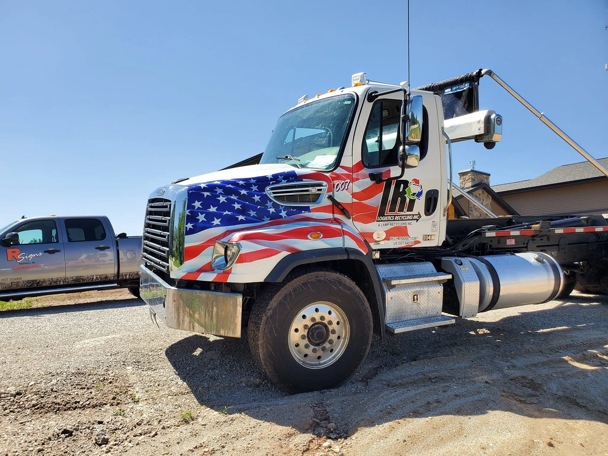 A flatbed truck decorated with an American flag design on its front. Parked on a gravel lot under a clear blue sky, with a silver pickup truck and a house in the background.