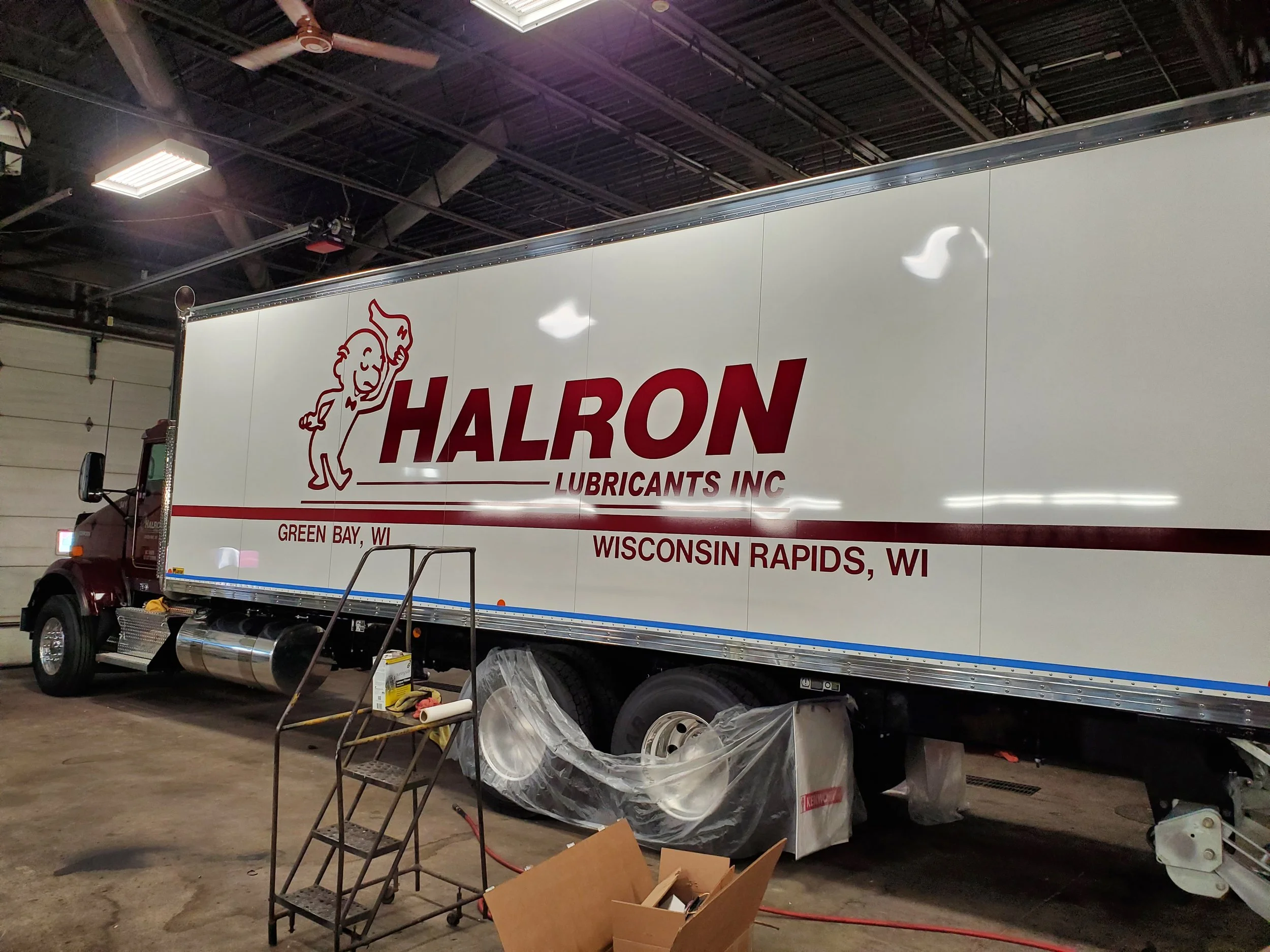 A large white truck with the logo of Halron Lubricants Inc., a smiling character giving a thumbs-up. The truck is parked inside a garage or warehouse. There are steps, tools, and plastic covering parts of the truck visible in the area.