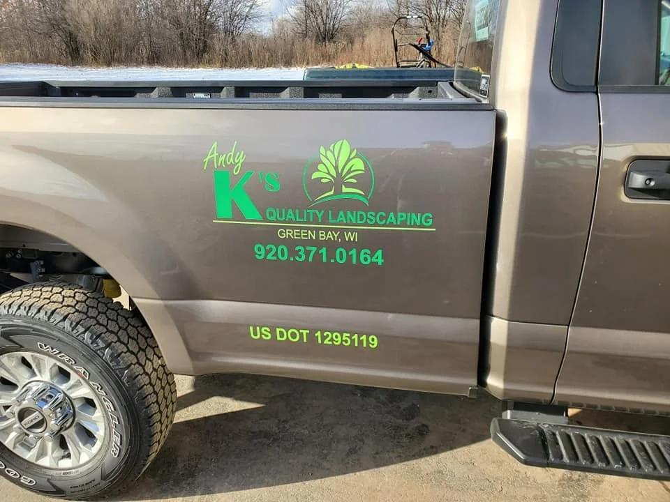 A gray pickup truck with green lettering and logo for Andy K's Quality Landscaping, located in Green Bay, Wisconsin, including a phone number and US DOT regulation number.