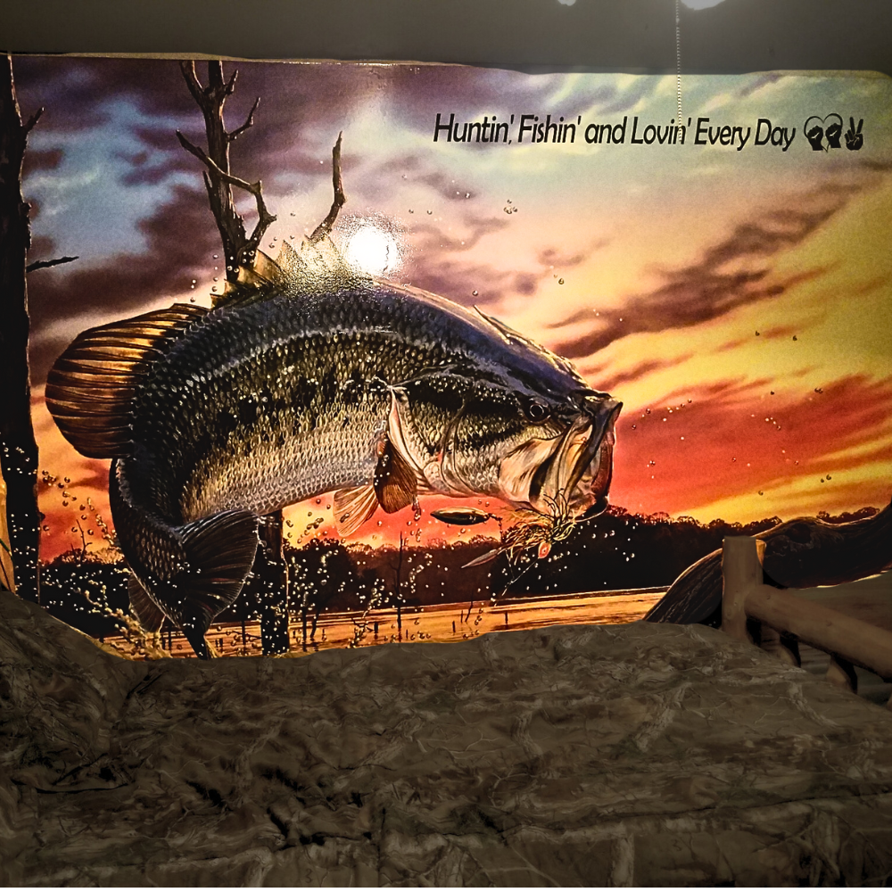 A large fish illustration with a sunset background, showing trees and a body of water, and the text "Huntin', Fishin' and Lovin' Every Day" with deer and peace sign icons.