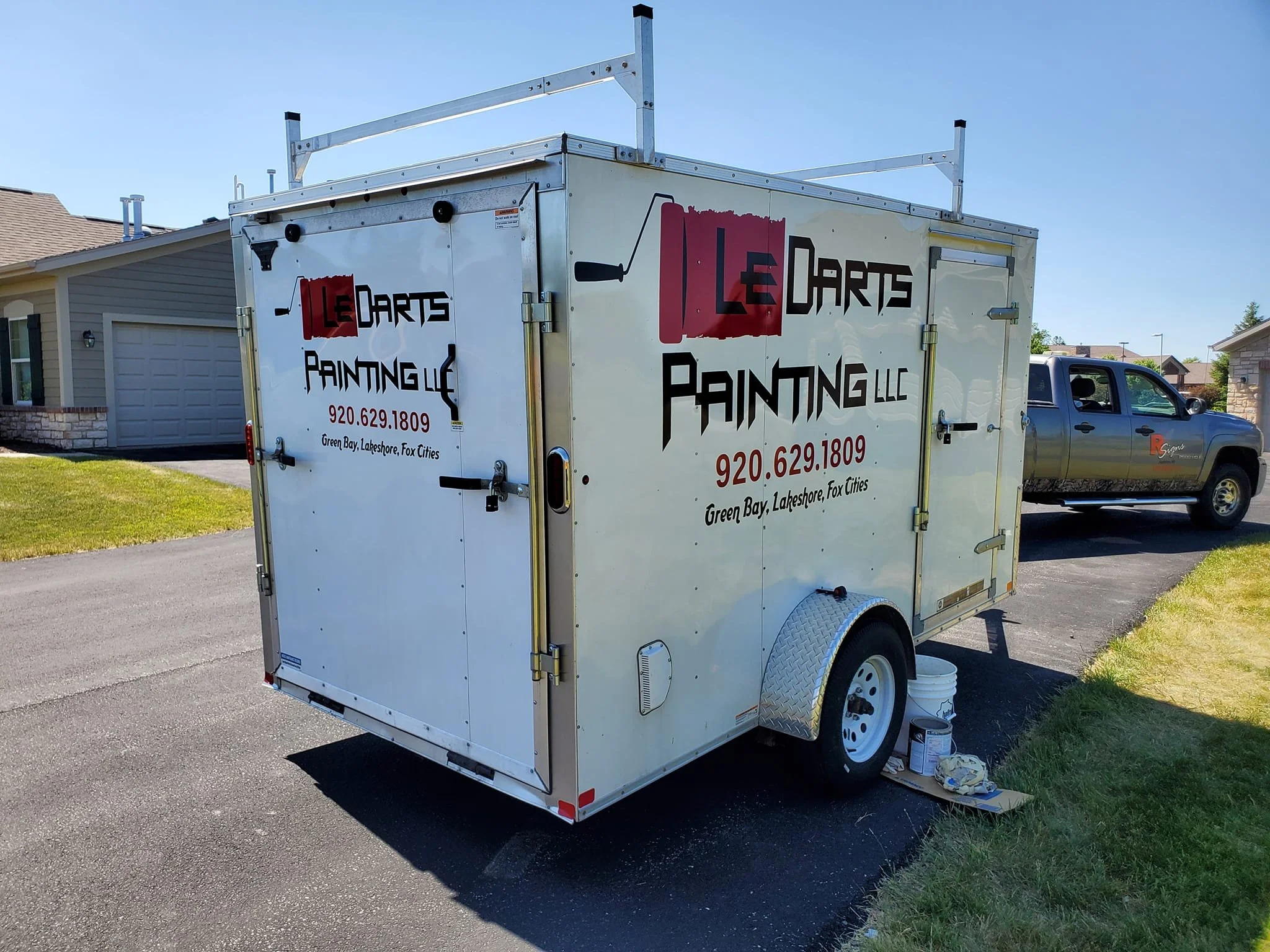 A mobile painting business trailer with company name "Le Darts Painting LLC" written on it, parked on a driveway next to a pickup truck, under a clear blue sky.