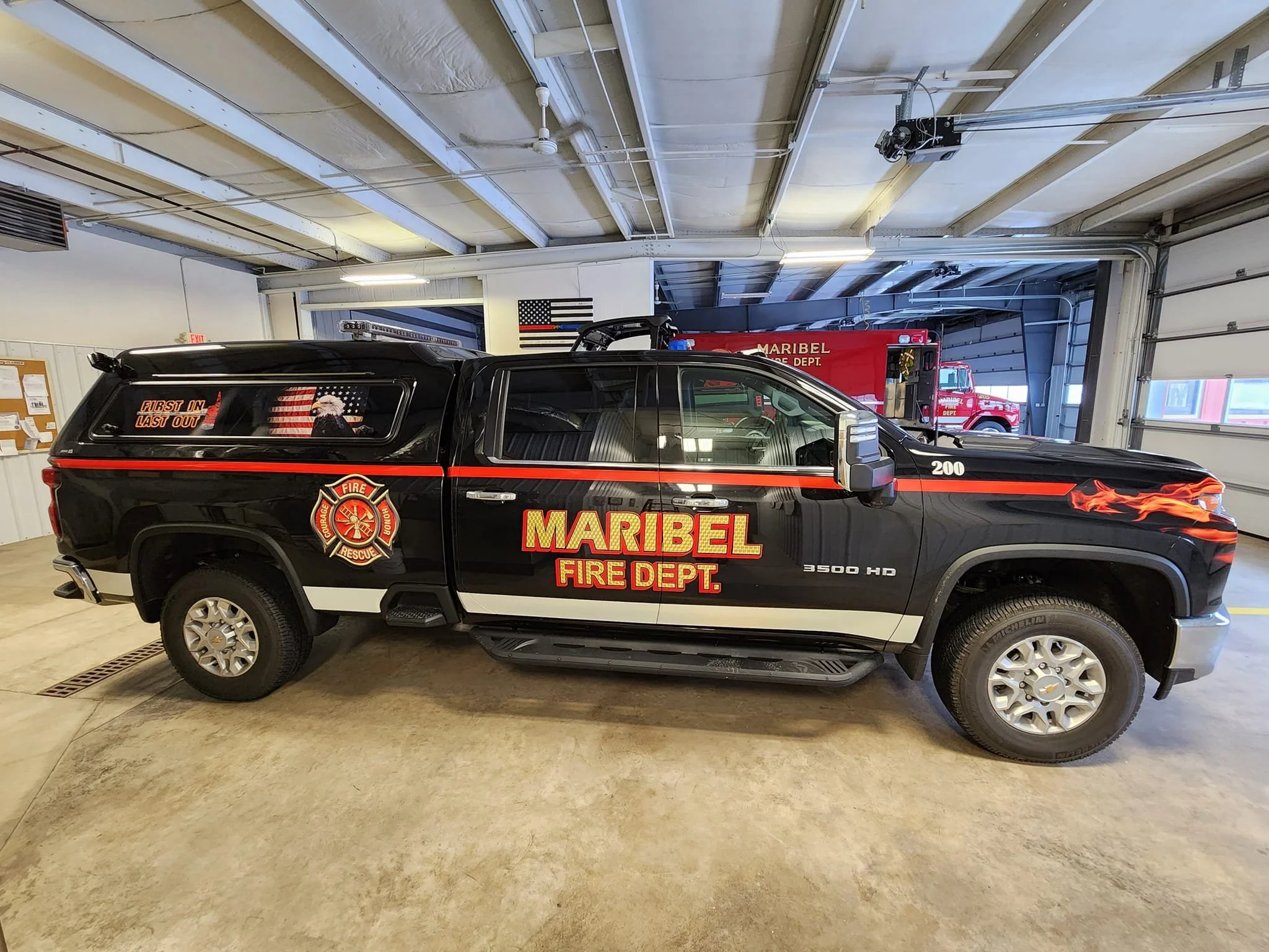 Black Maribel Fire Department pickup truck with red and yellow decals and flames on the front, parked inside a fire station with a red fire truck in the background.