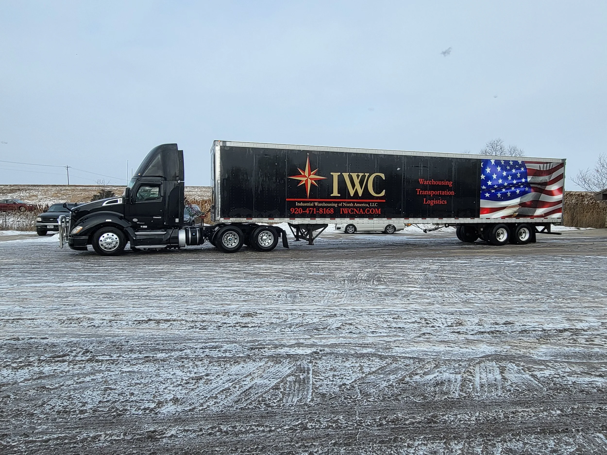 Black semi-truck with a trailer displaying the logo and branding of IWC, Industrial Warehousing of North America, LLC, on a snow-covered lot with a few parked cars and trees in the background.