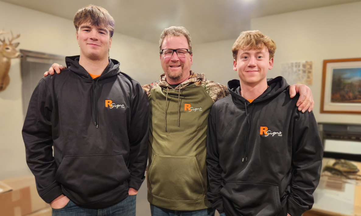 Three men posing together indoors, smiling, with two young men wearing black hoodies with orange logos on either side of an older man wearing a camouflage-patterned hoodie and glasses.