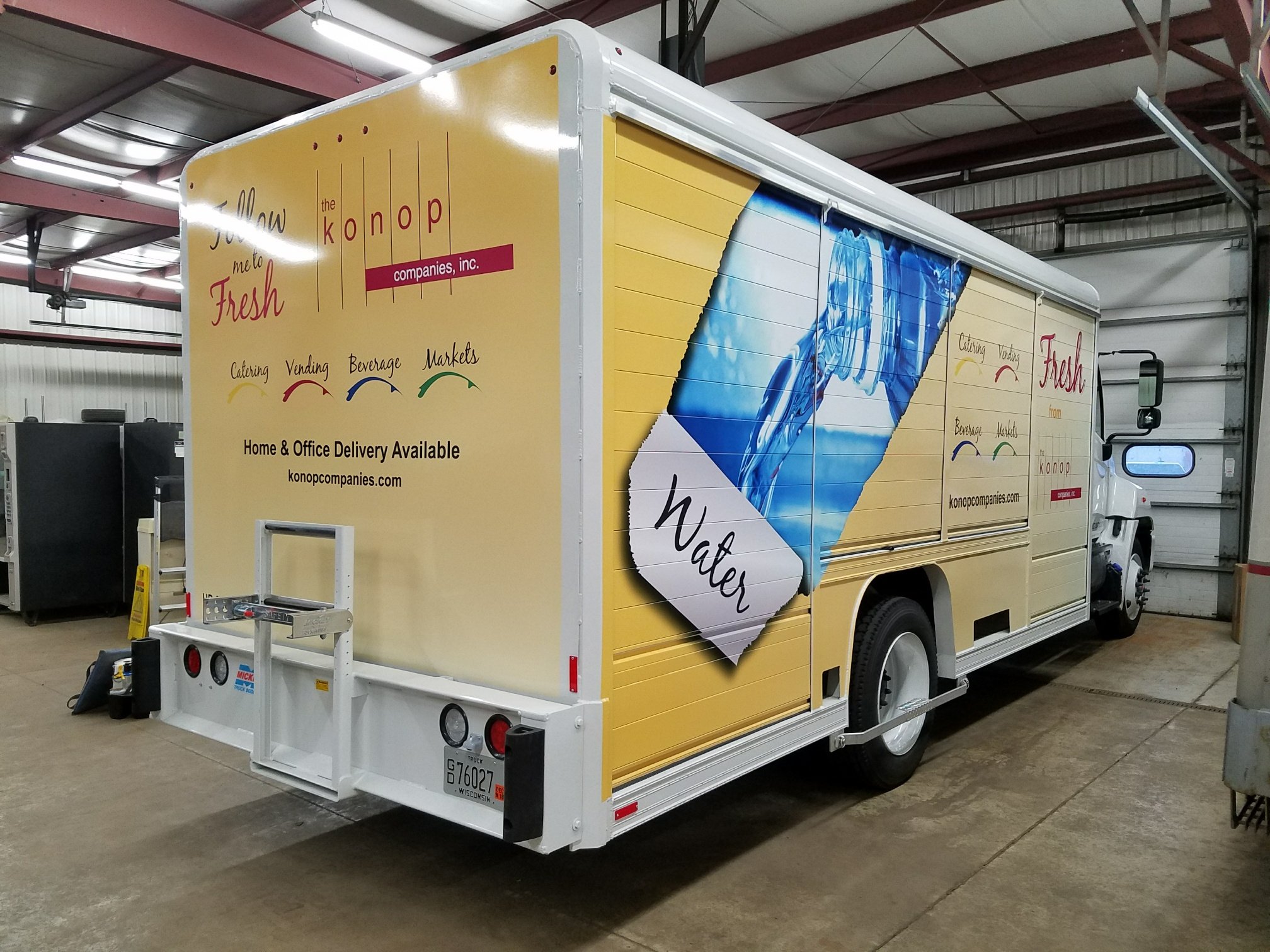 A delivery truck with advertisements for 'The Konop Companies, Inc.', showcasing water bottles, cleaning supplies, and beverages, parked in an indoor warehouse.
