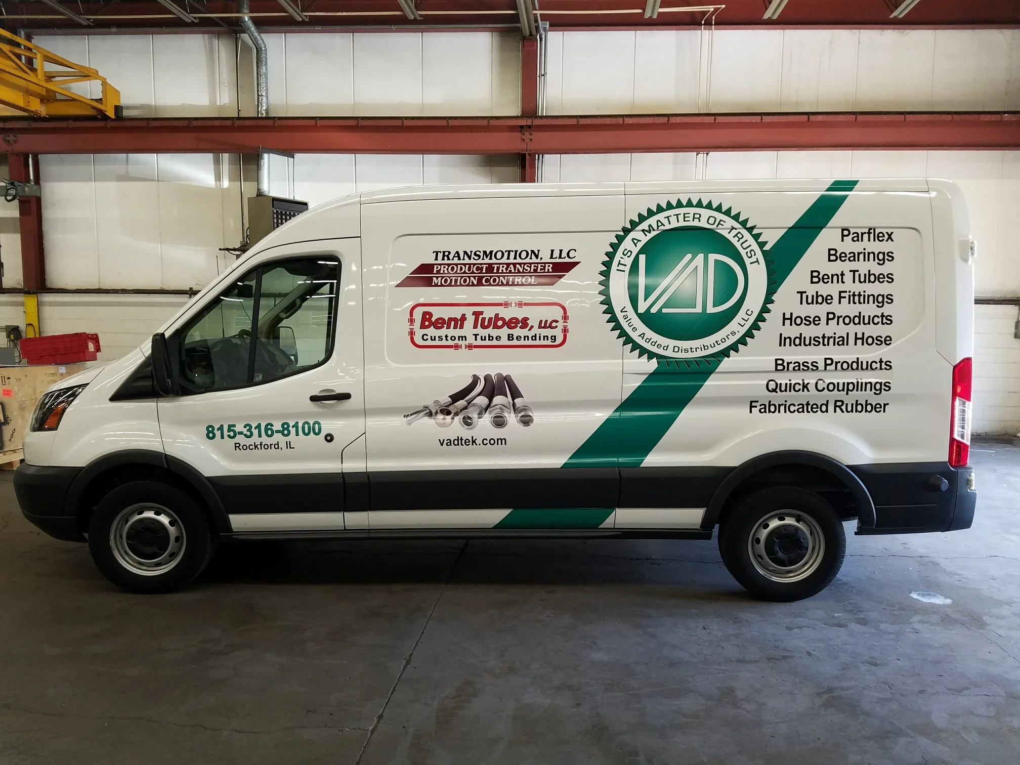 Side view of a white delivery van with green, black, and red graphics and text advertising a company that sells industrial hoses, fittings, and brass products, parked inside a warehouse.