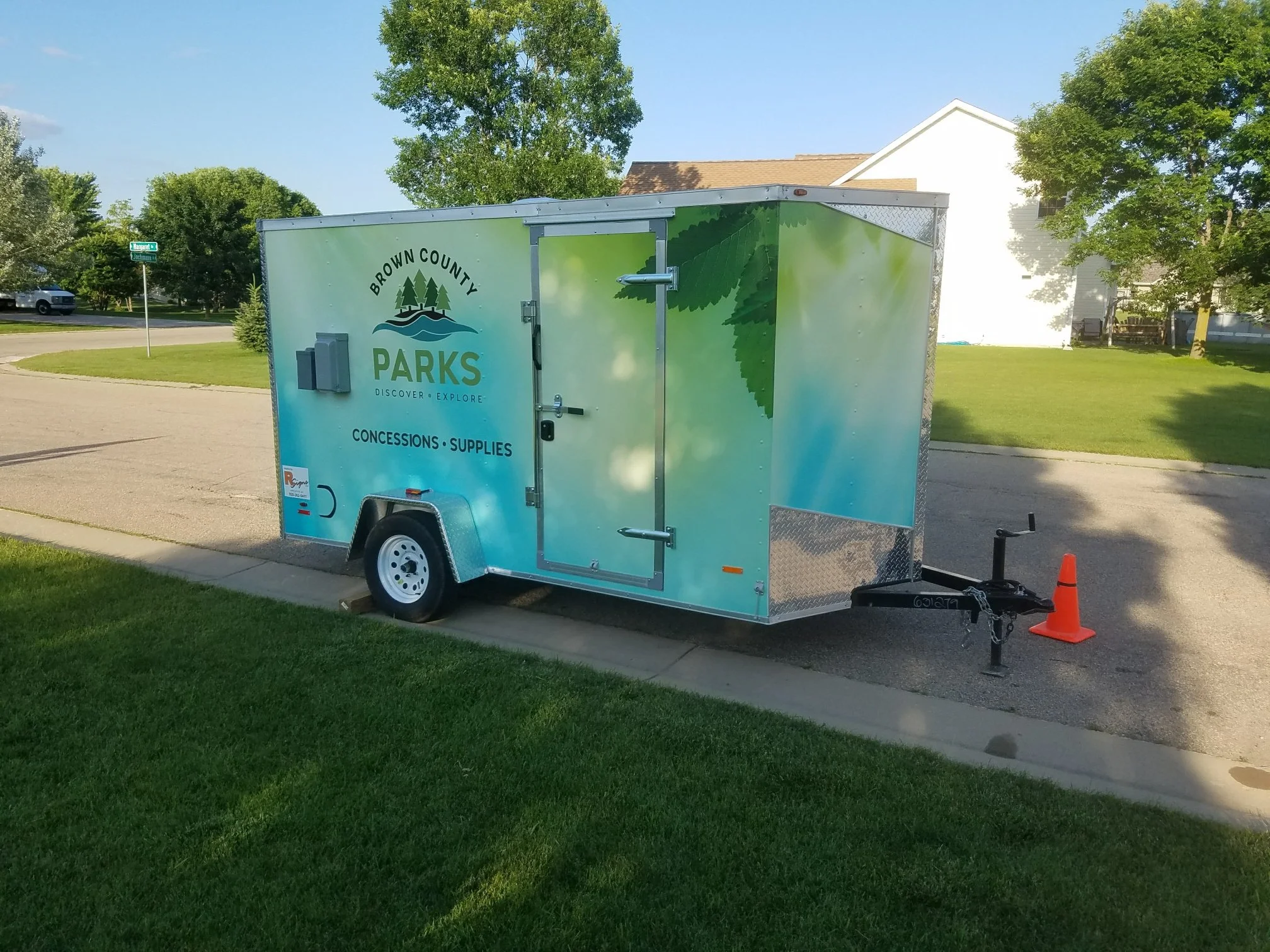A travel trailer for Brown County Parks, parked on a residential street. The trailer is blue with green tree graphics, and text indicating it offers concessions and supplies. There is an orange traffic cone next to the trailer's hitch.