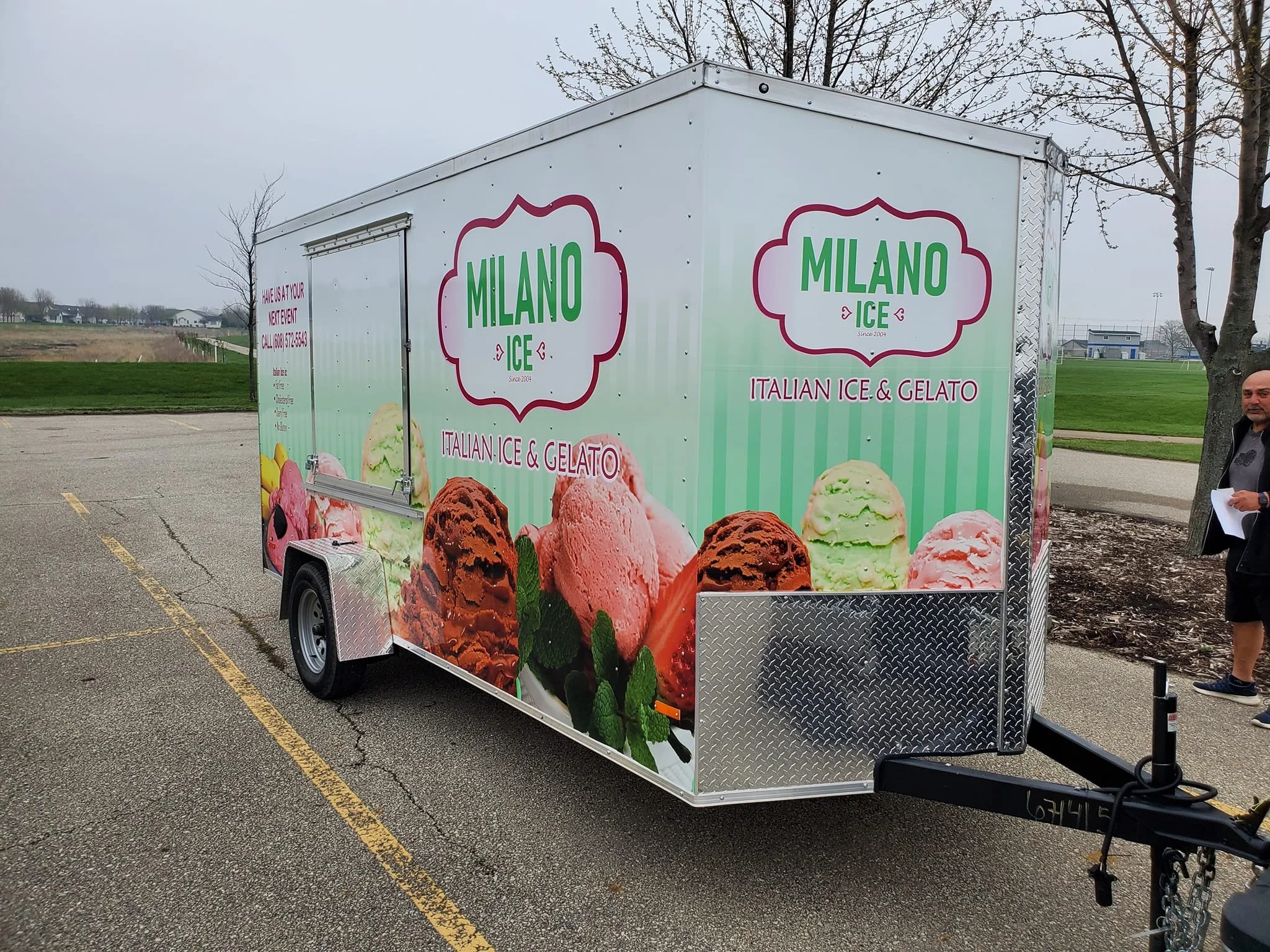 Ice cream truck with Milano Ice branding, displaying images of various ice cream flavors, parked in an outdoor lot on a cloudy day.