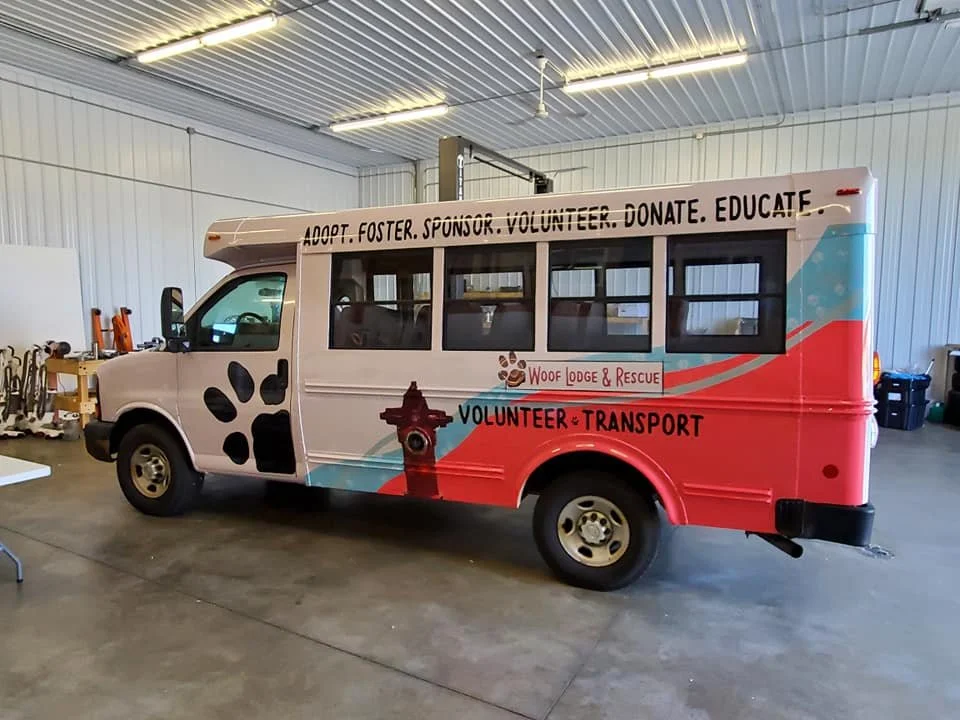A small bus wrapped in a colorful decal promoting dog rescue, adoption, volunteering, and education, parked inside a warehouse or large garage.
