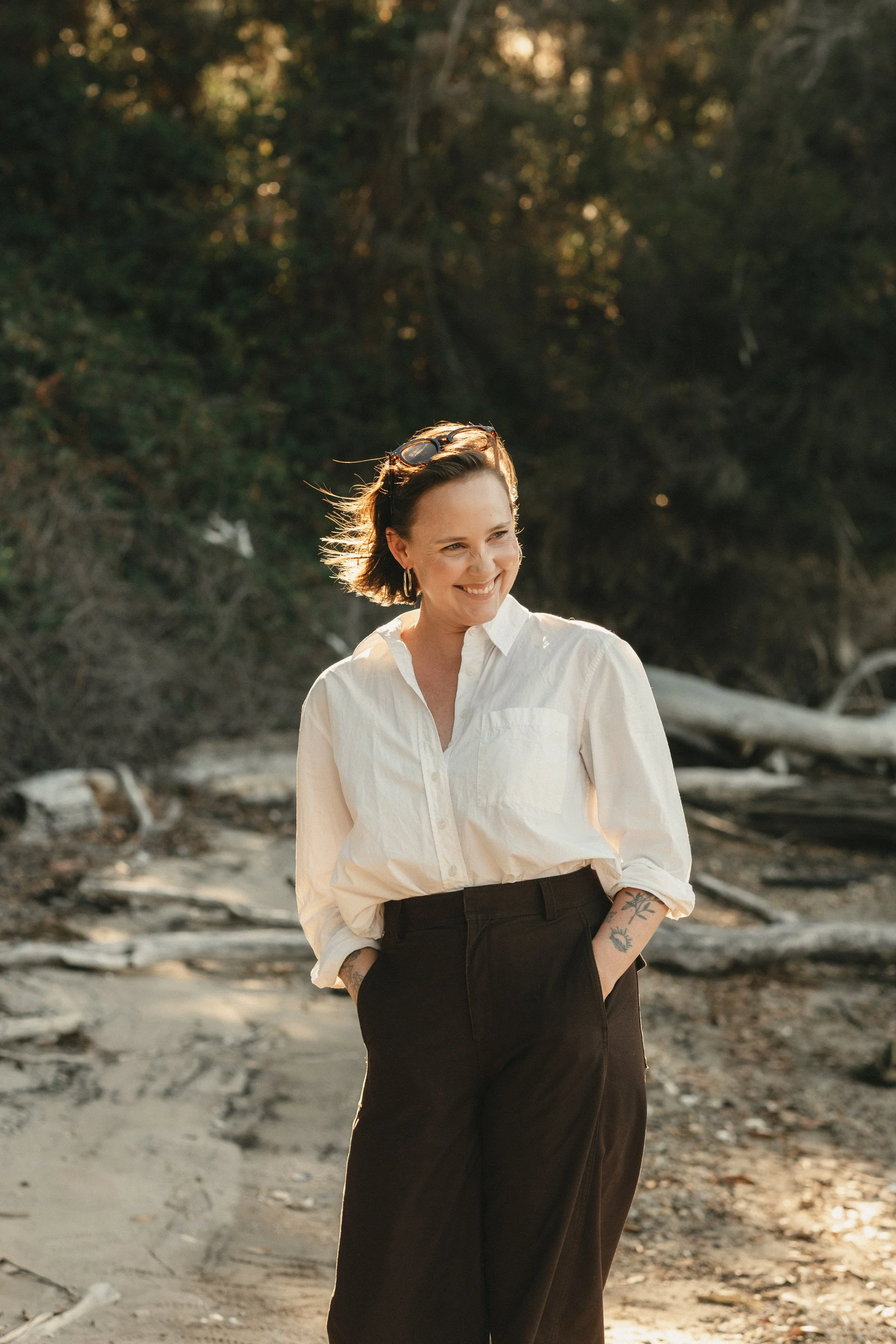 A woman smiling outdoors on a sandy and rocky beach, with trees and driftwood in the background.