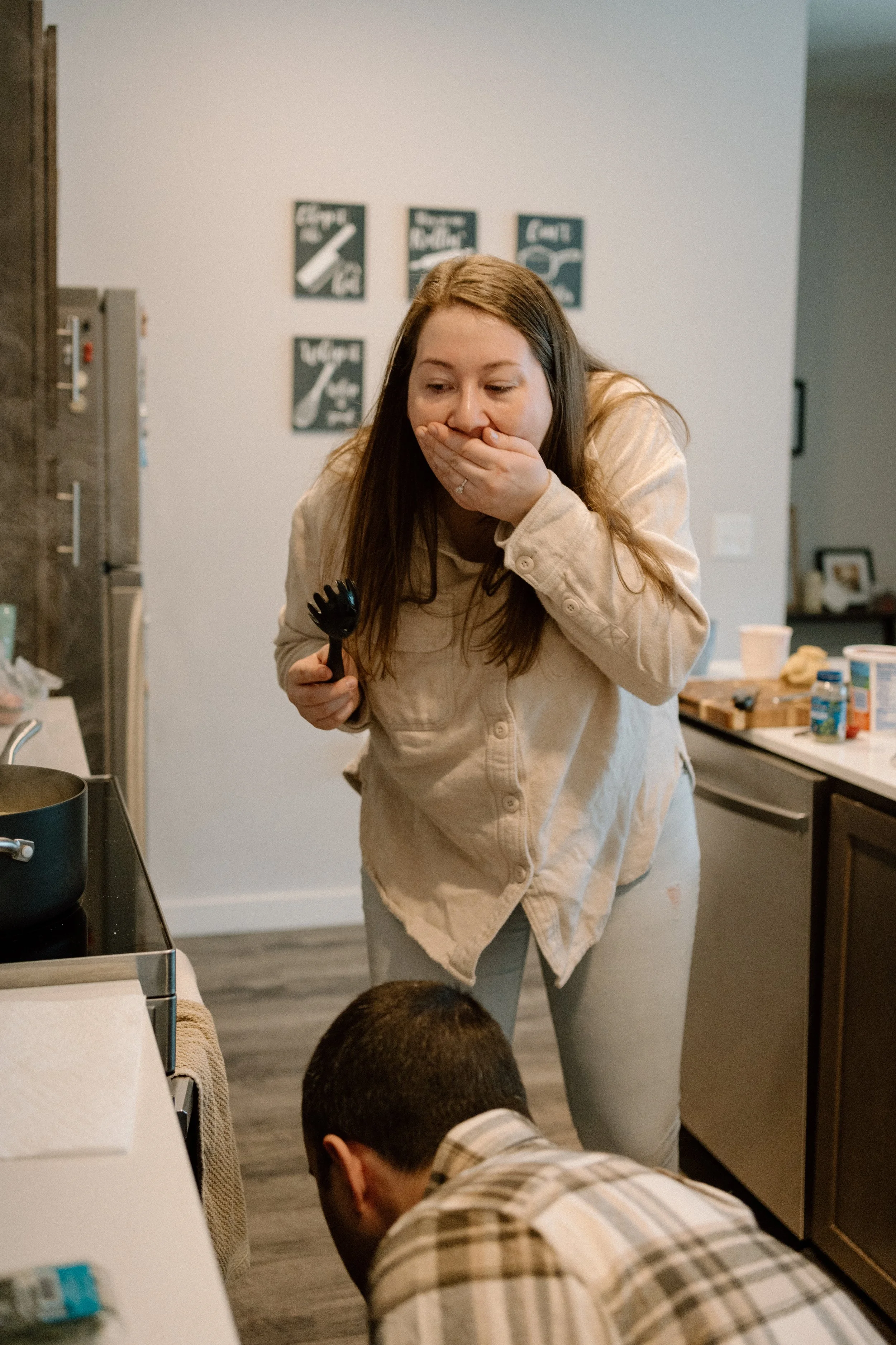 A woman in a beige jacket covering her mouth with her hand, standing in a kitchen, holding a black kitchen utensil, looking down at a man who is bent over nearby.