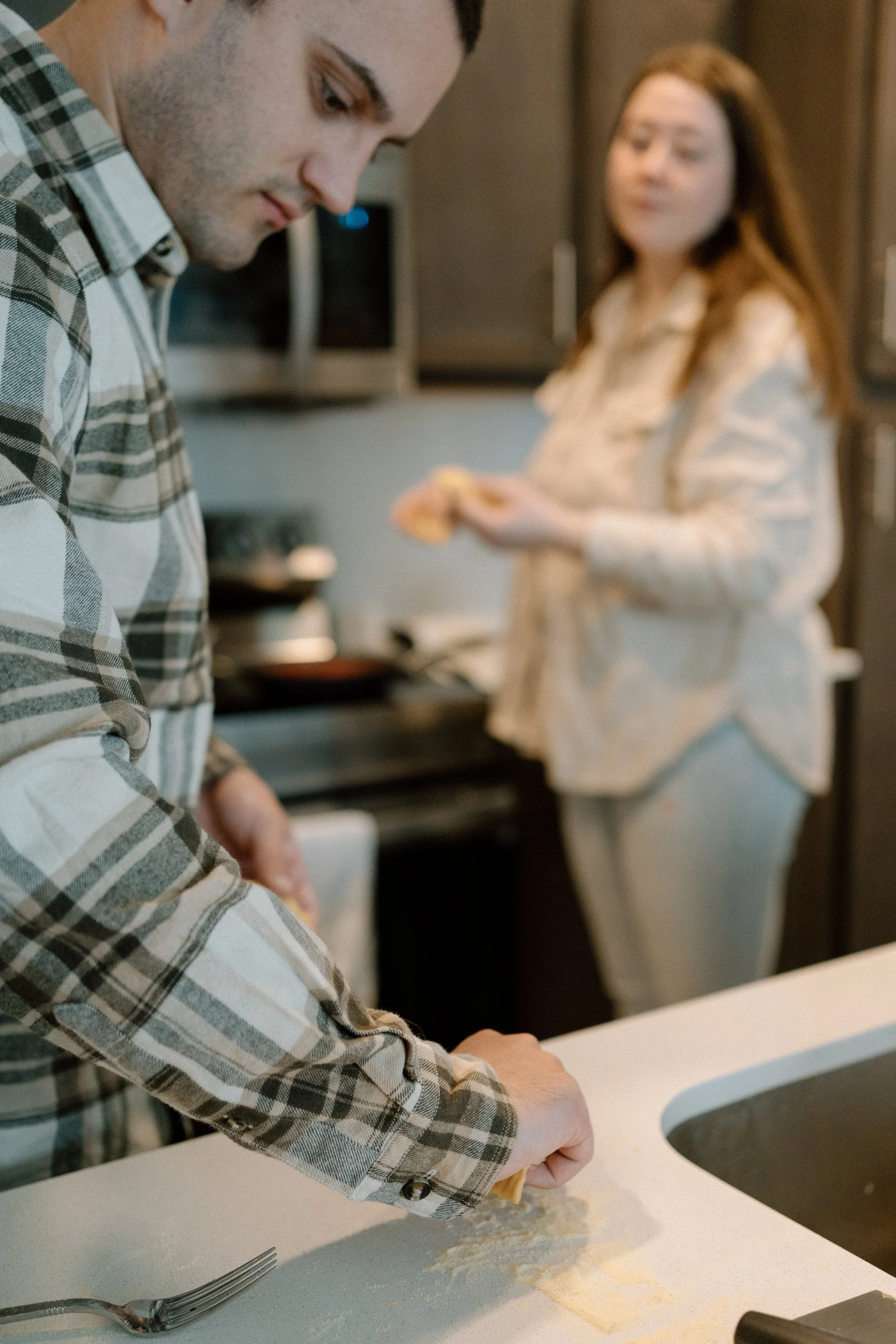 A man in a plaid shirt is rolling out dough on a kitchen countertop, with a woman in the background holding a piece of dough or bread, looking at him.