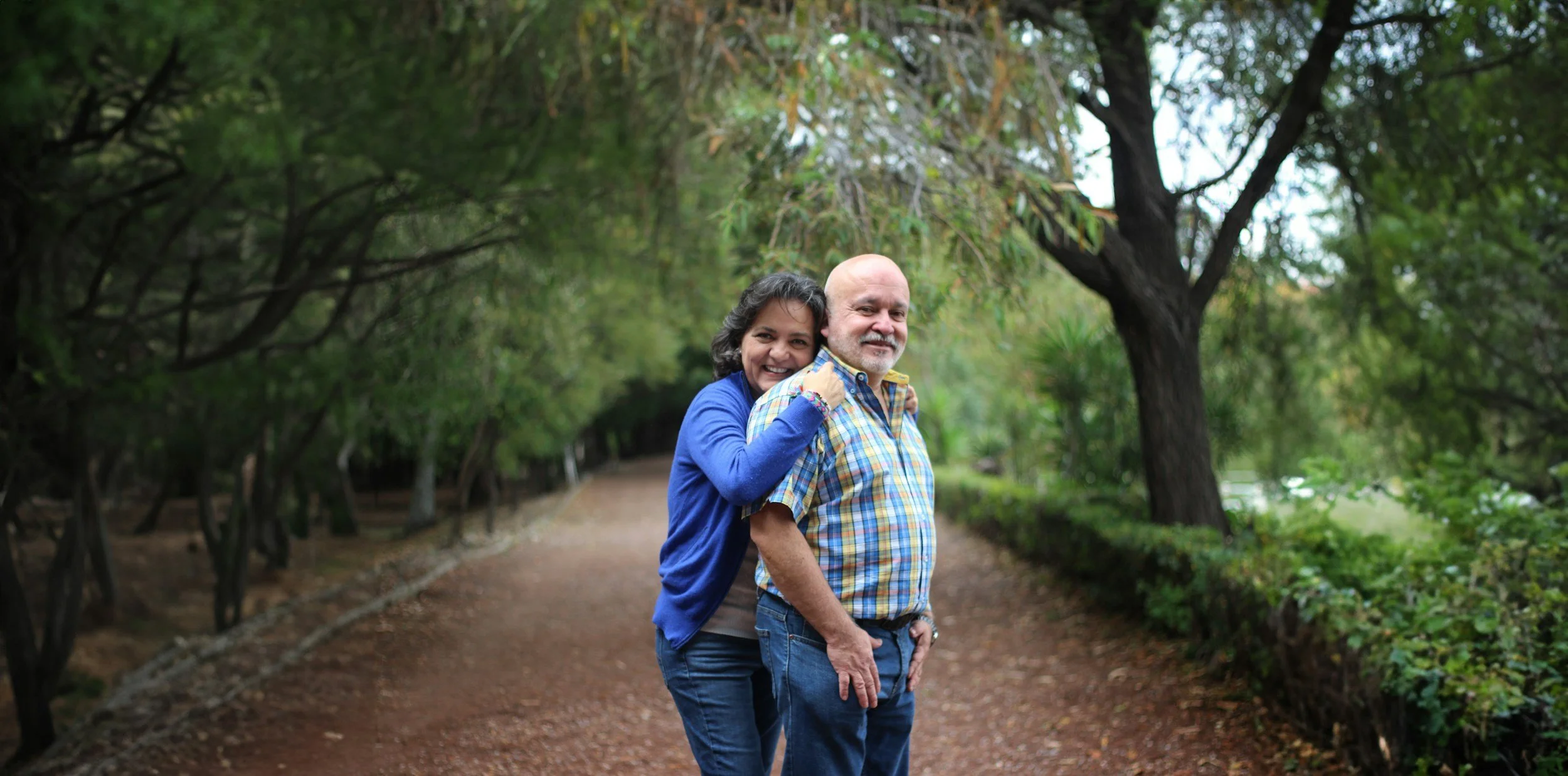 A couple stands on a wooded park trail, smiling, with the woman hugging the man from behind and the man in a plaid shirt looking at the camera, surrounded by green trees and foliage.