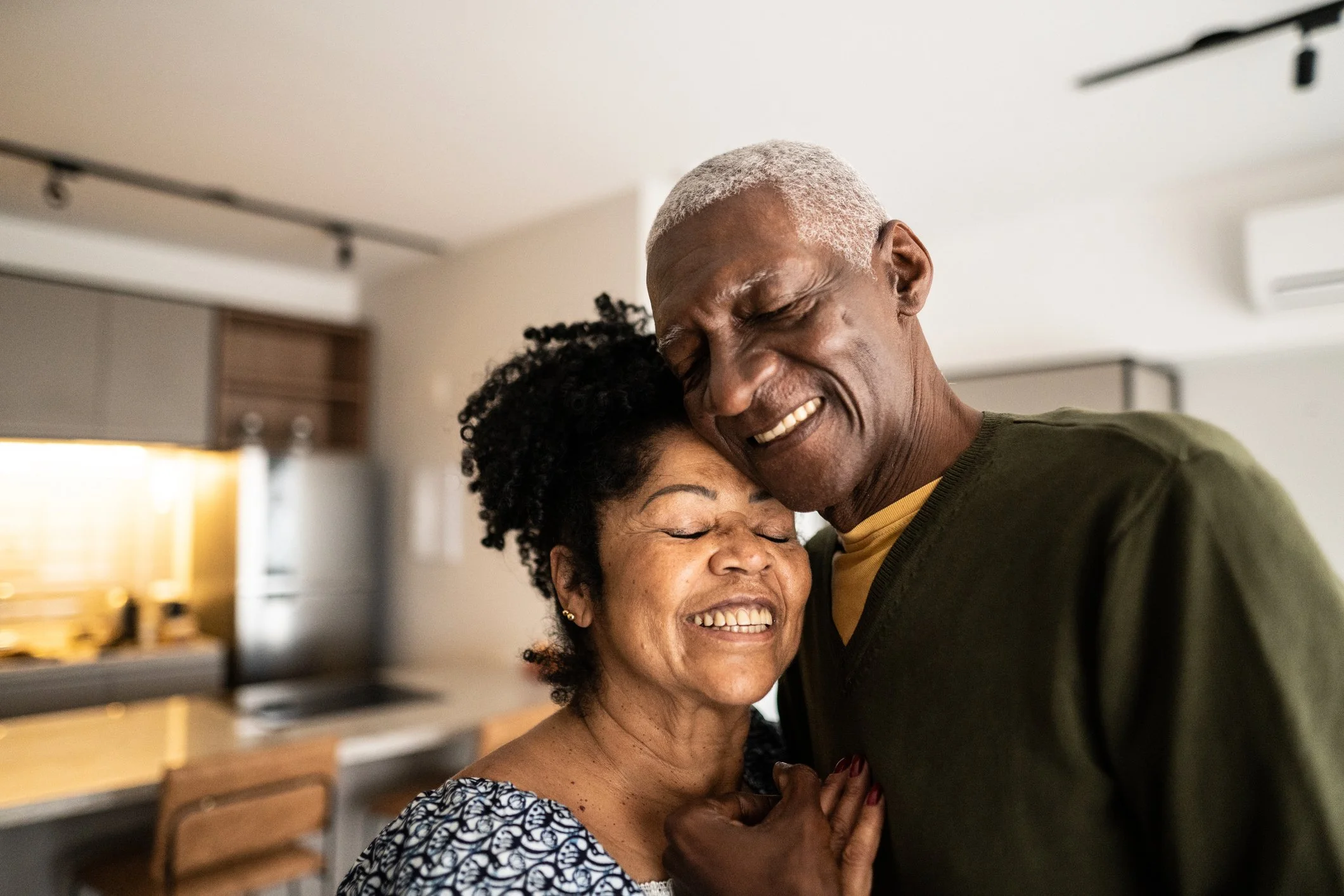 A joyful elderly couple embraces in a cozy kitchen, both smiling with their eyes closed, expressing love and happiness.