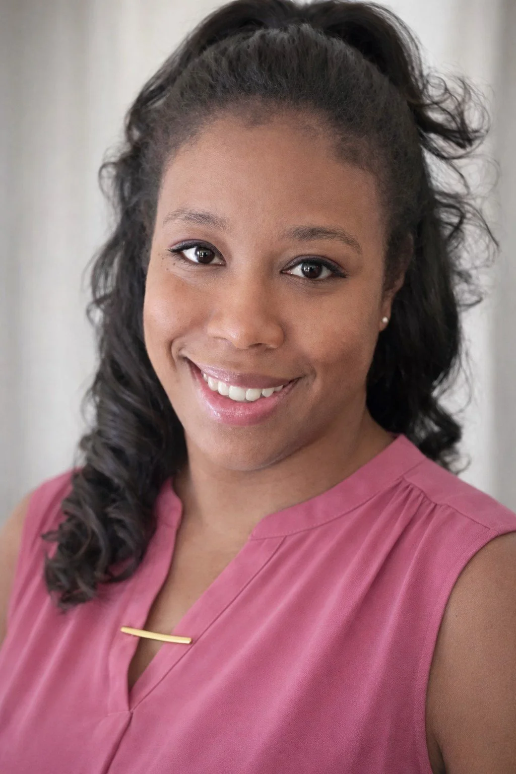 A smiling woman with dark, curly hair tied back, wearing a pink sleeveless blouse and small pearl earrings.