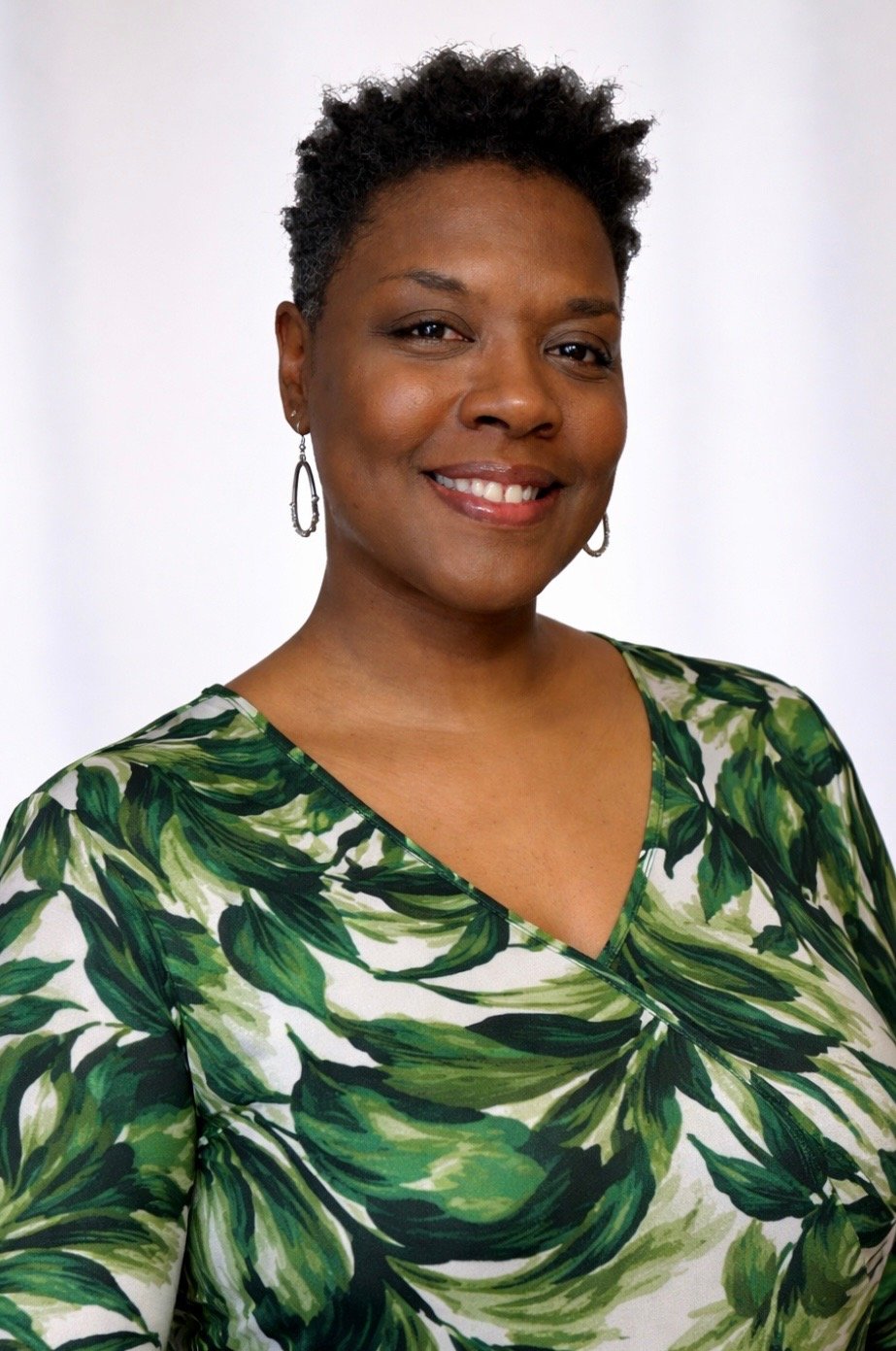 A woman with short curly hair smiling, wearing a green and white leaf-patterned blouse and silver earrings, against a plain white background.