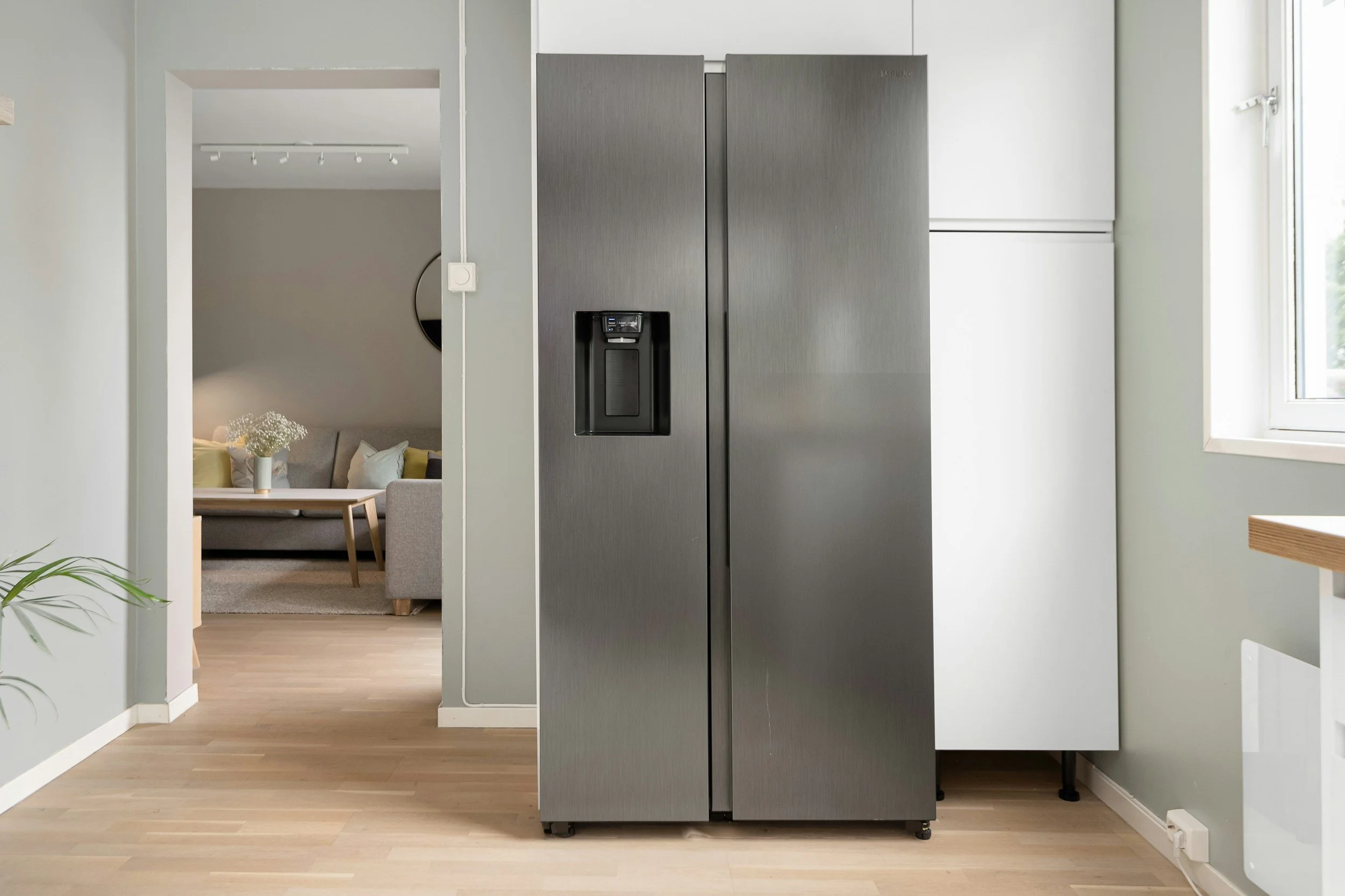 Stainless steel side-by-side refrigerator in a kitchen, with a living room visible through an open doorway