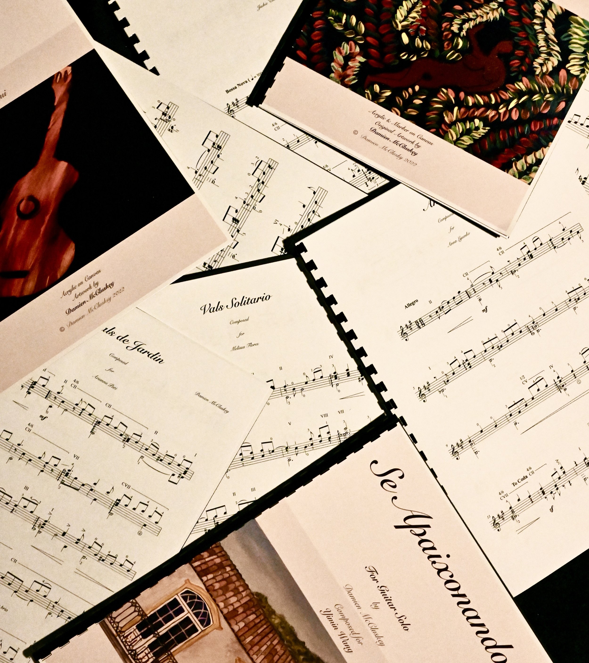 Collection of music sheets and artwork pages spread out on a surface, featuring handwritten musical notations and colorful images of floral embroidery and a wooden object.