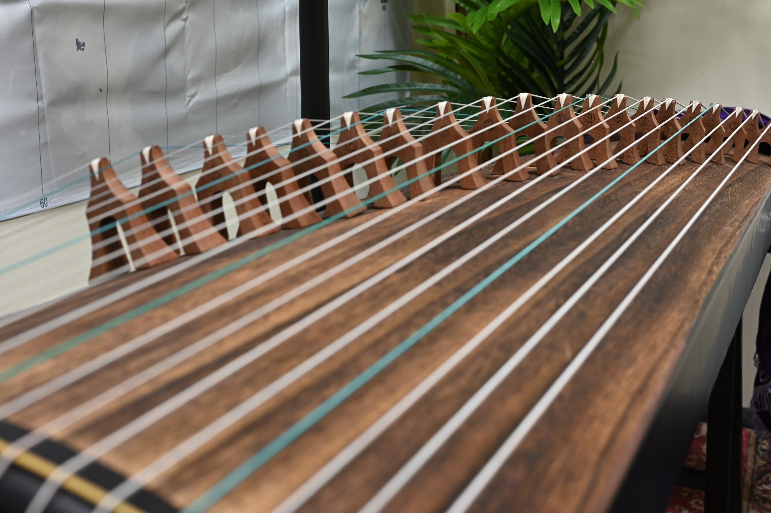 A wooden stringed musical instrument, likely a hammered dulcimer, with metal strings stretched over a wooden frame and tuning pegs.
