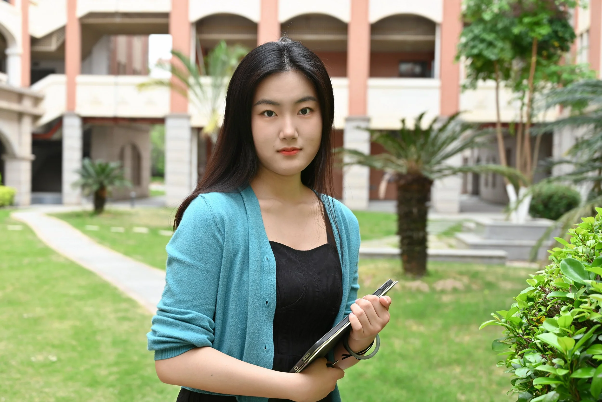 Young woman with long dark hair wearing a blue cardigan and black top, holding a tablet, standing outdoors in a garden with grass, trees, and a building in the background.
