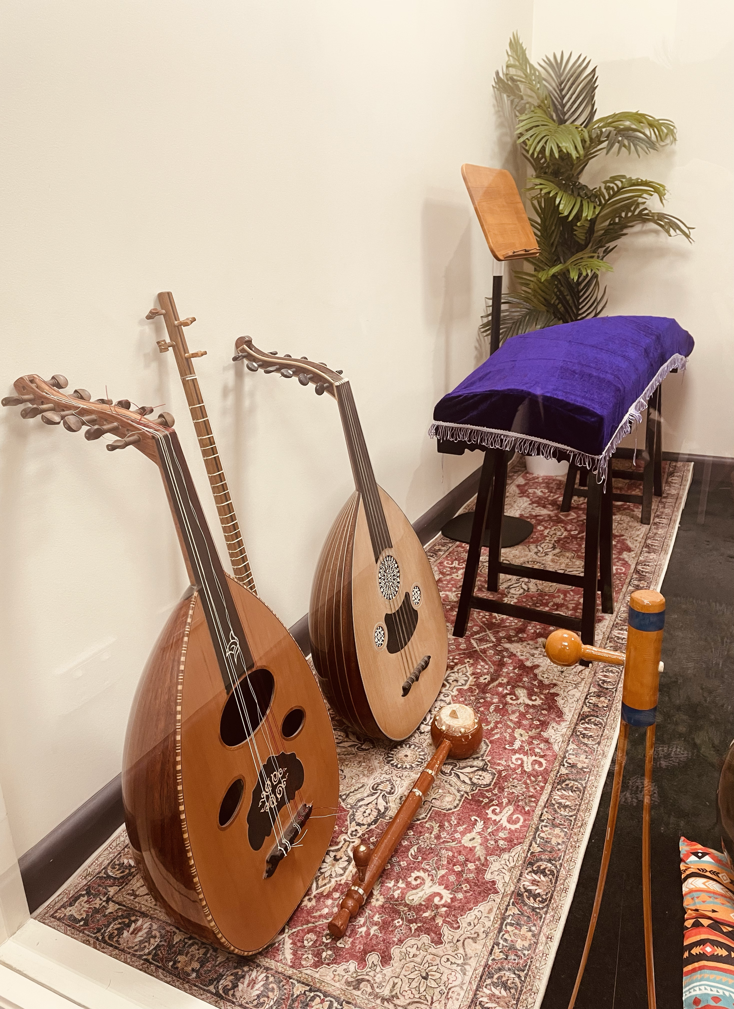 Two stringed musical instruments, likely ouds, resting on a patterned rug, next to a black stool covered with a purple cloth, a stand with a wooden music holder, and a large potted plant in the background.