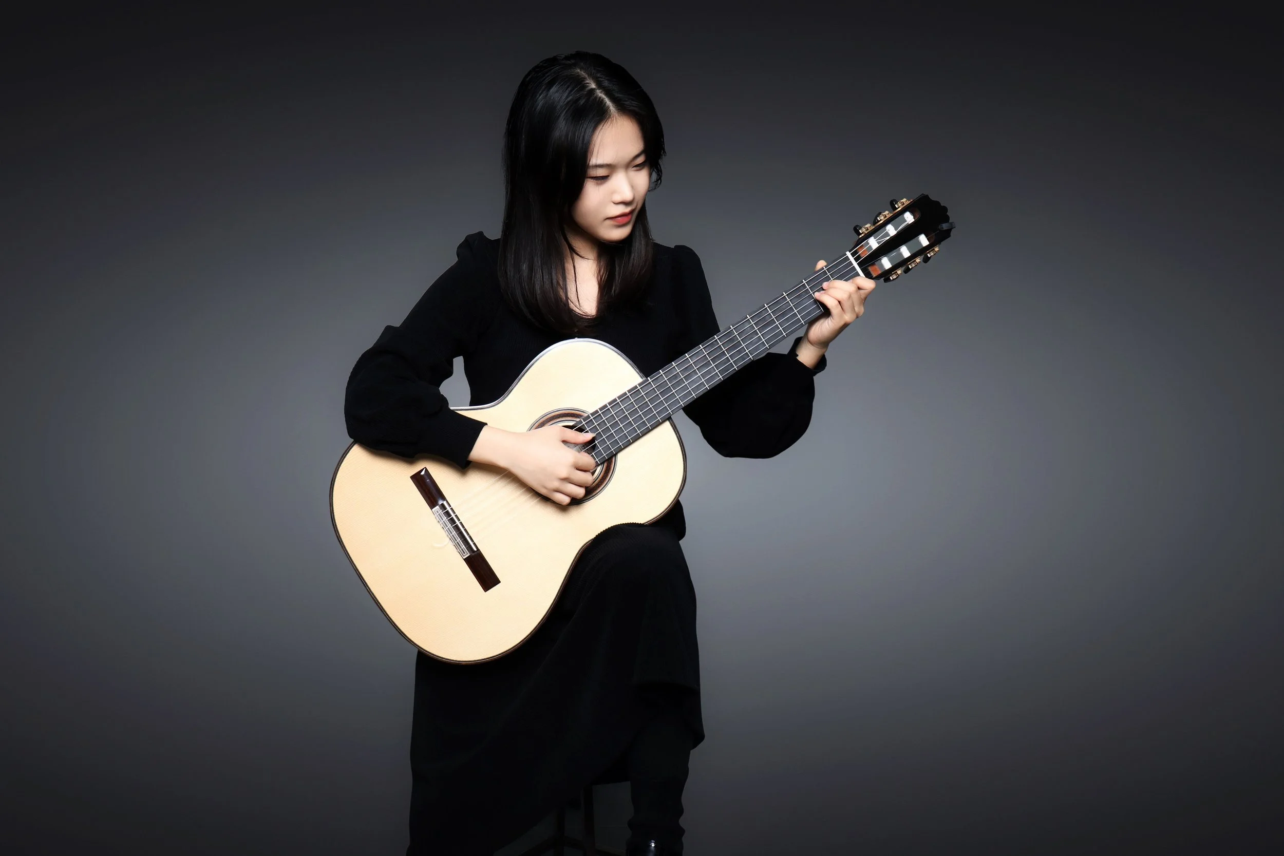 Young woman sitting on a stool playing an acoustic guitar against a dark background.