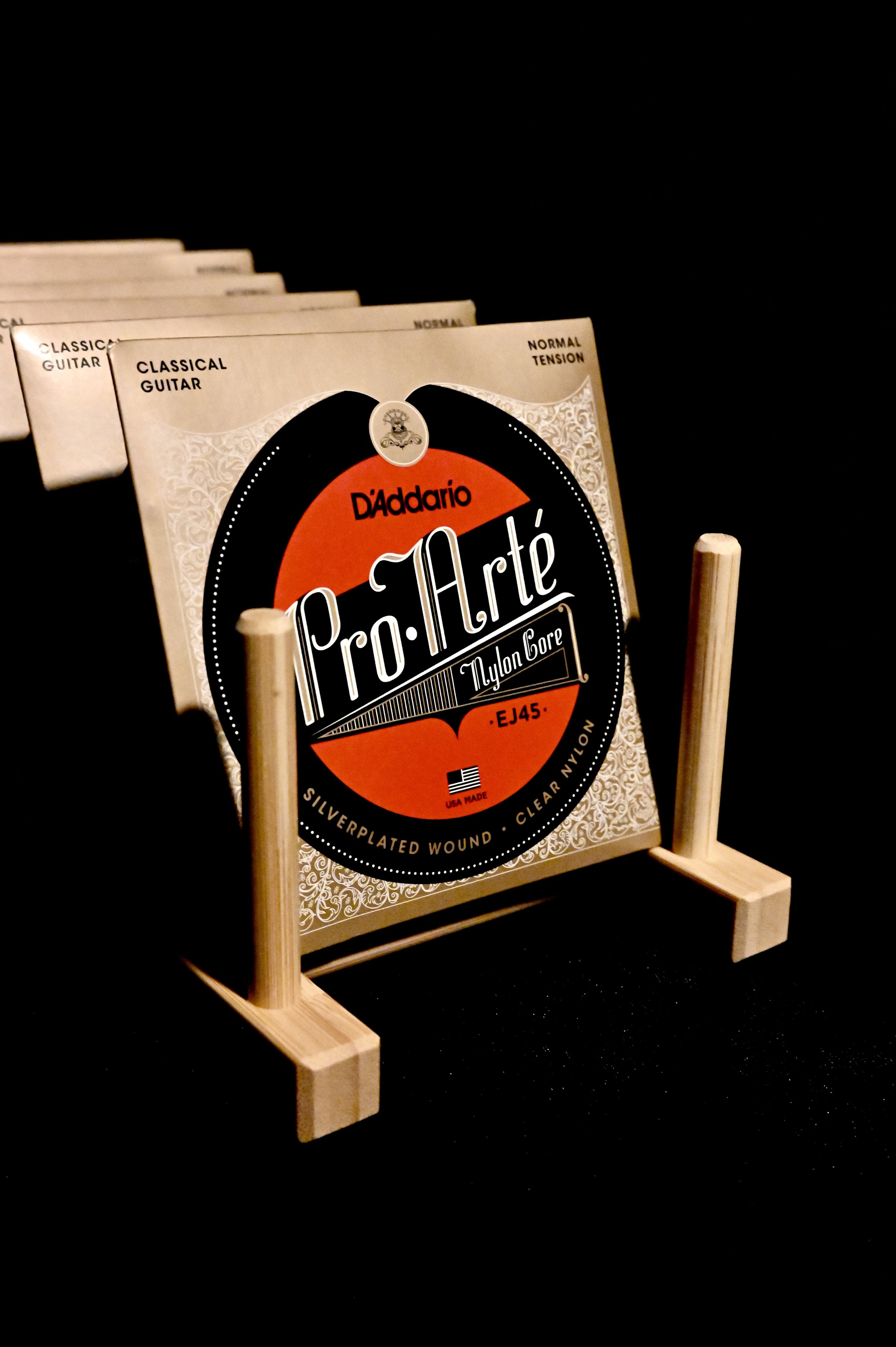 A pack of classical guitar strings on a small wooden display stand, with several more packs behind it, against a black background.