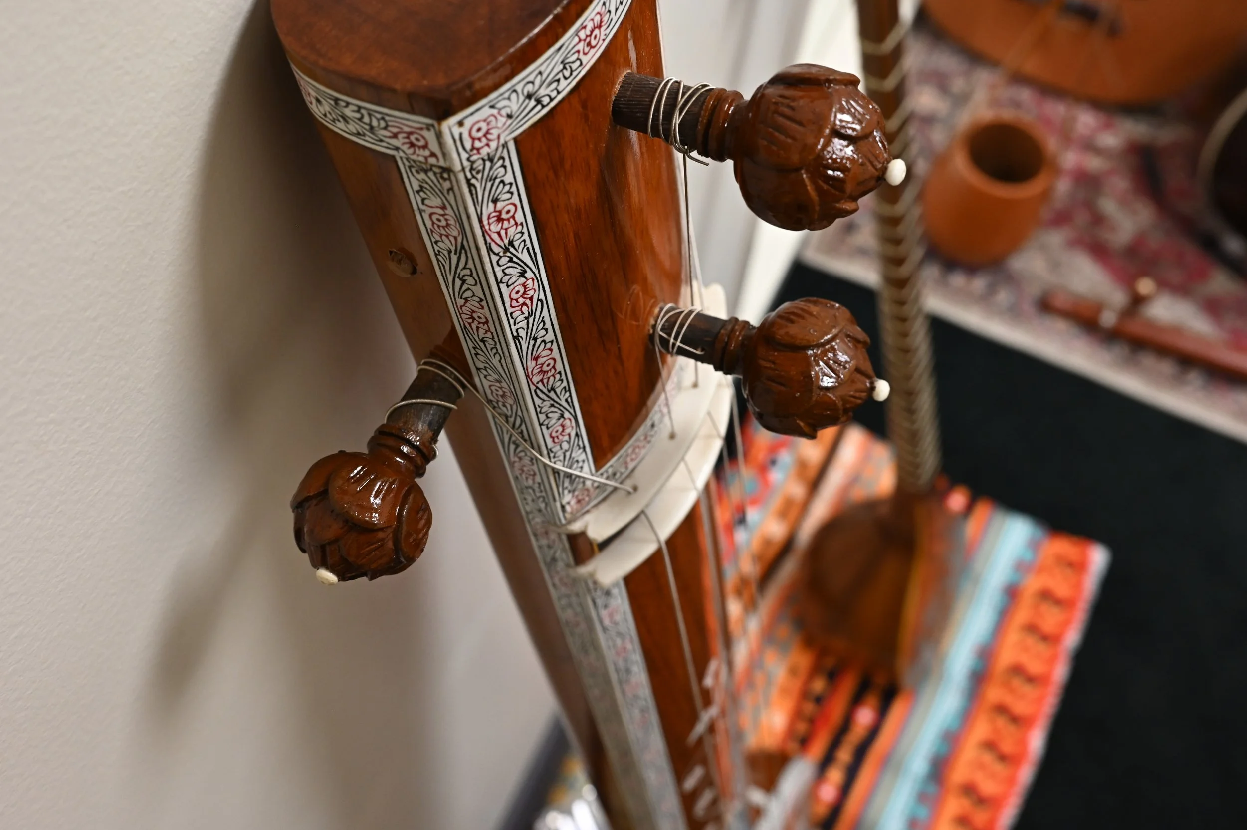 Close-up of a stringed instrument's headstock with ornate wooden tuning pegs resembling carved faces, on a wooden body with decorative inlay, against a background of rugs and pottery.