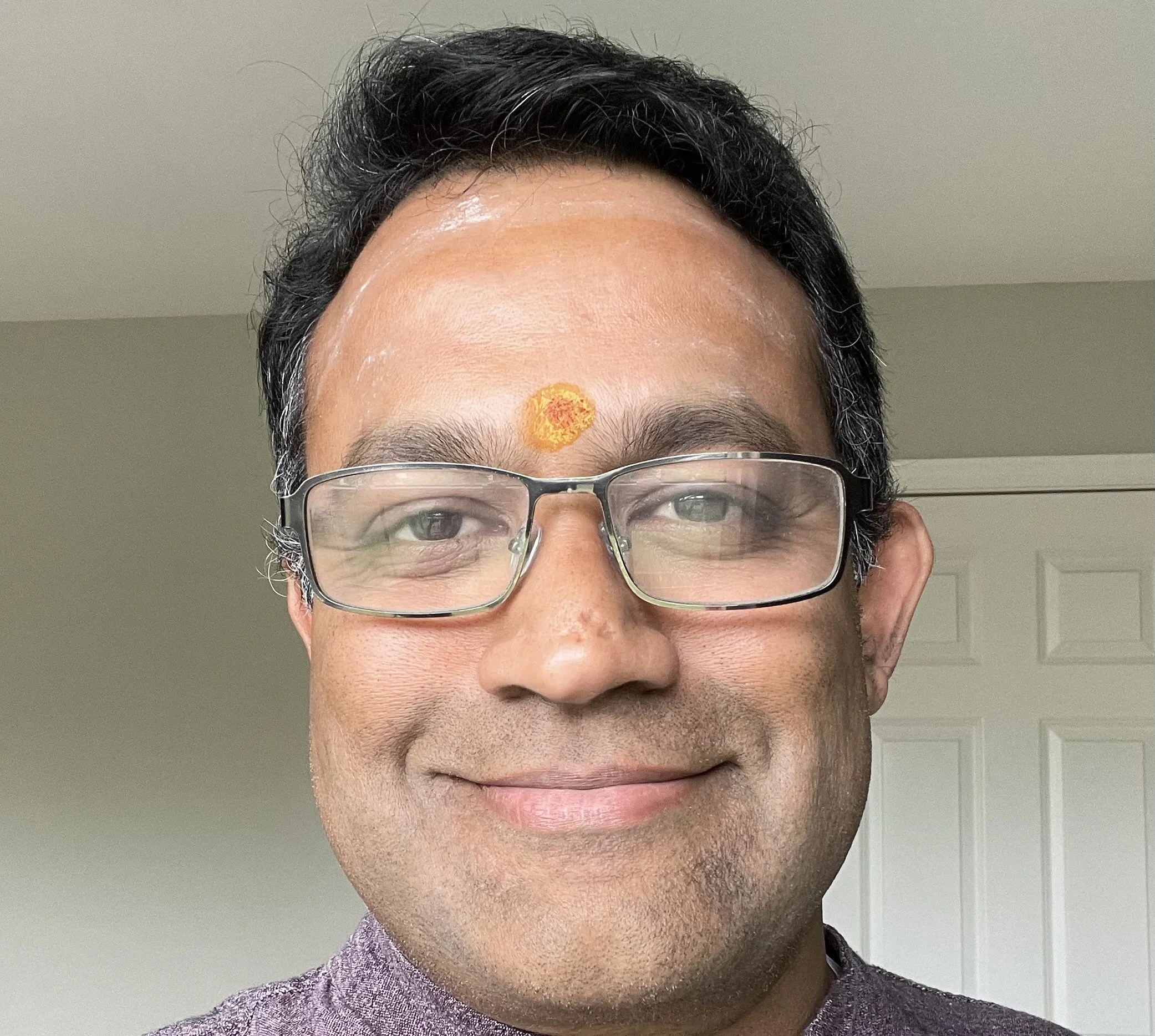 A man smiling, wearing glasses and a purple shirt, with a yellow and orange mark on his forehead, standing indoors in front of a light-colored wall and white door.