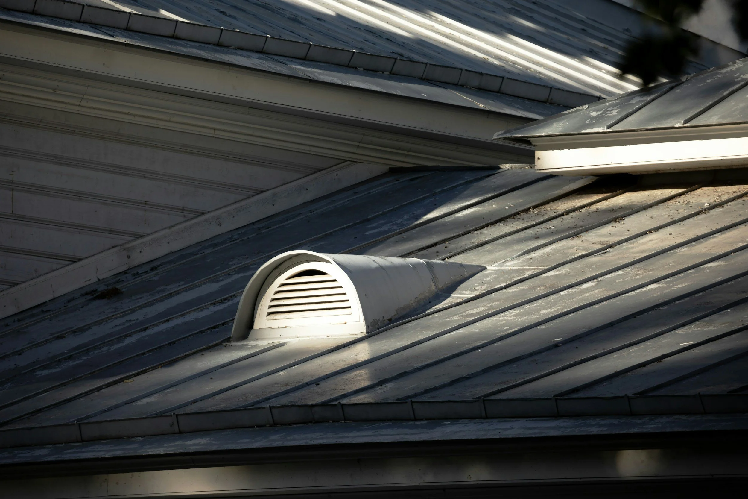 Close-up of a metal roof with a white vent pipe and flashing, showing overlapping panels and a weathered surface.
