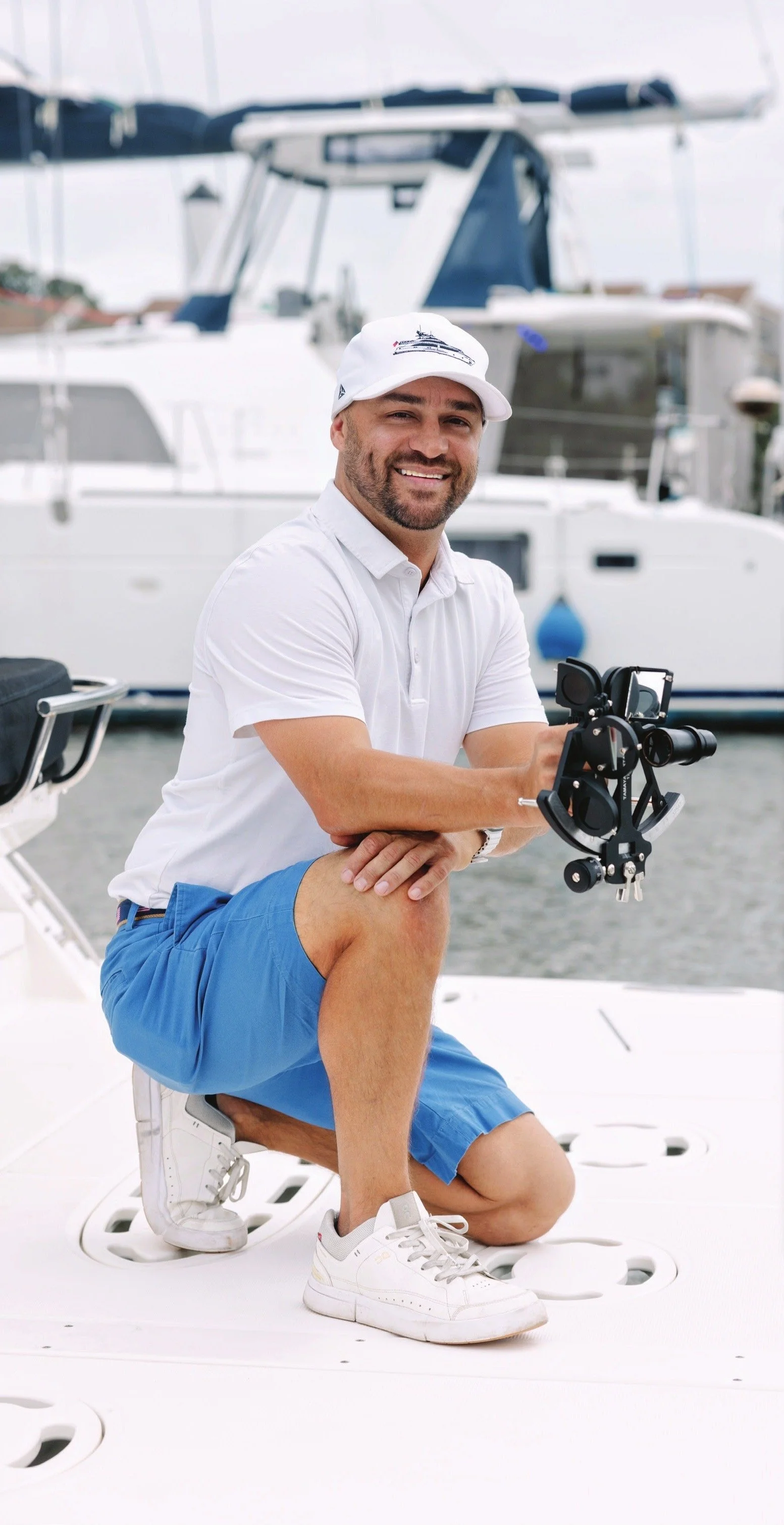 A man kneeling on a yacht deck holding a gimbal stabilizer, with a marina and large yacht in the background. He is smiling, wearing a white polo shirt, blue shorts, white sneakers, and a white cap.
