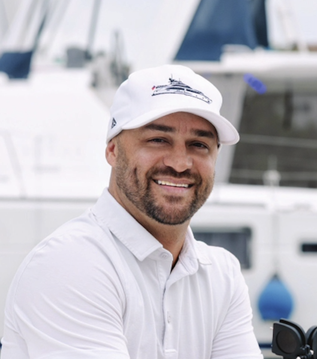 A captain smiling, wearing a white polo shirt and white cap with a boat logo, standing in front of yachts.