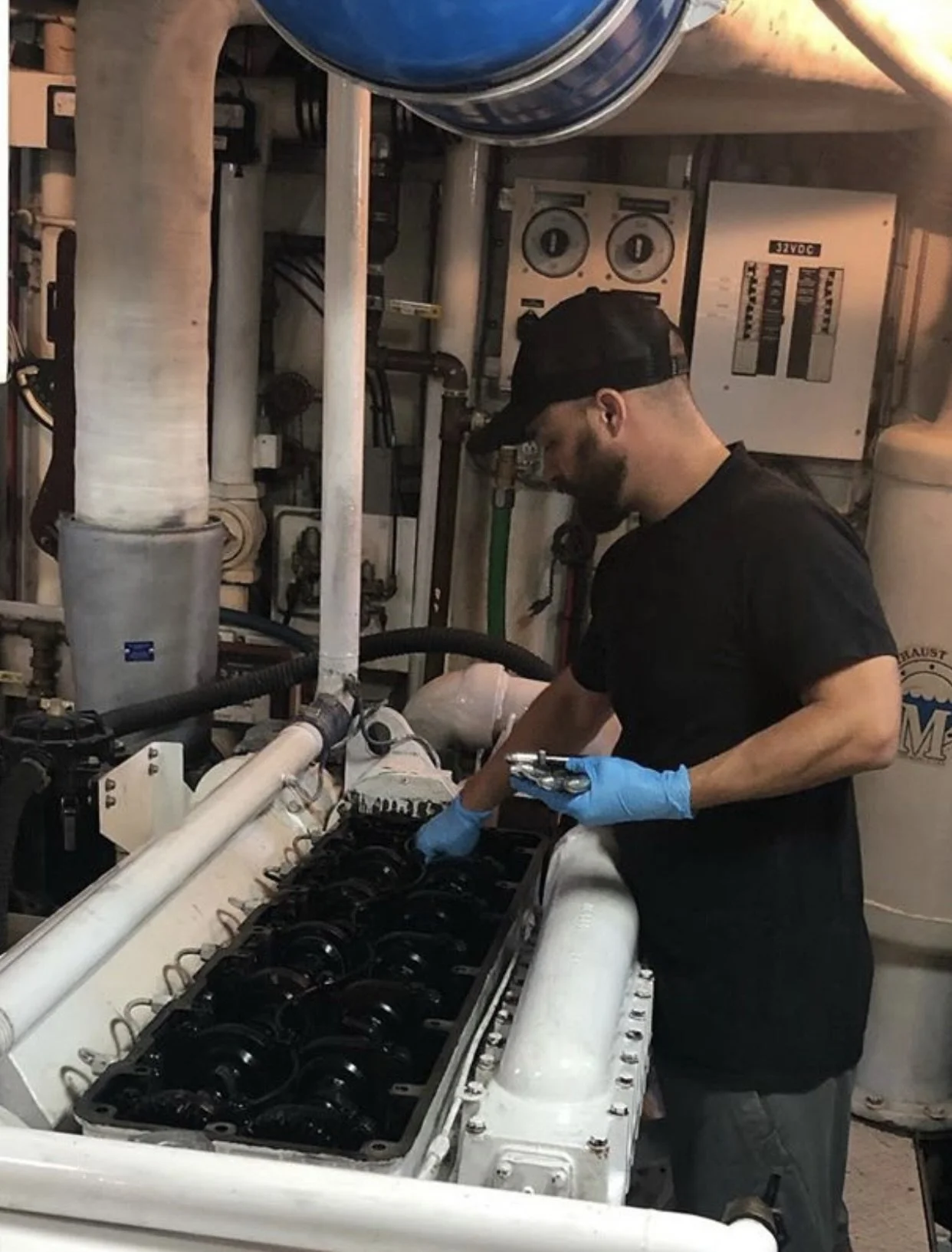 A man wearing a black cap, black t-shirt, and blue gloves working on a large mechanical or engine component in an industrial setting with pipes and electrical panels in the background.
